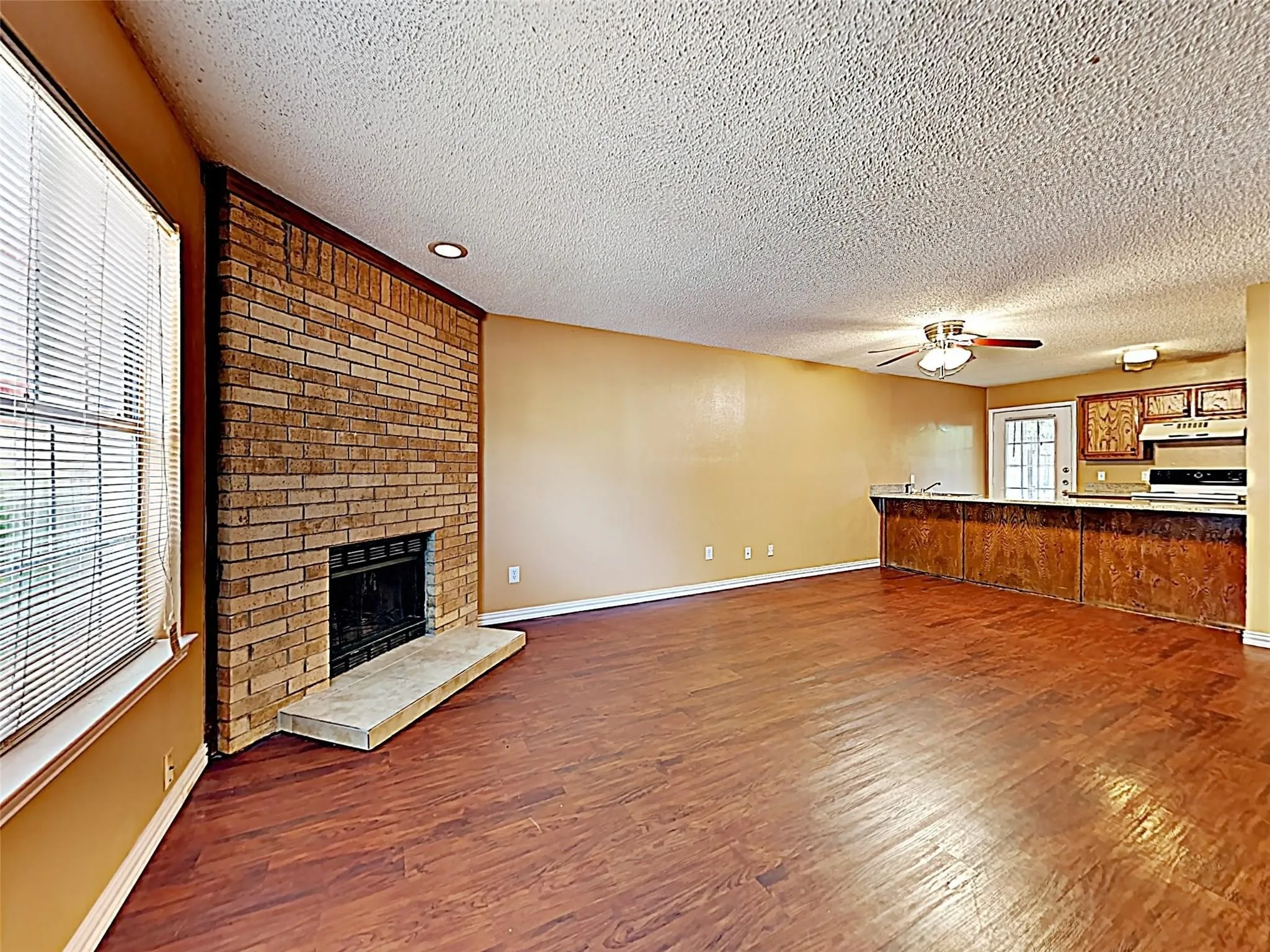 Living area featuring wood-finish flooring, a brick fireplace with hearth, and a window with blinds