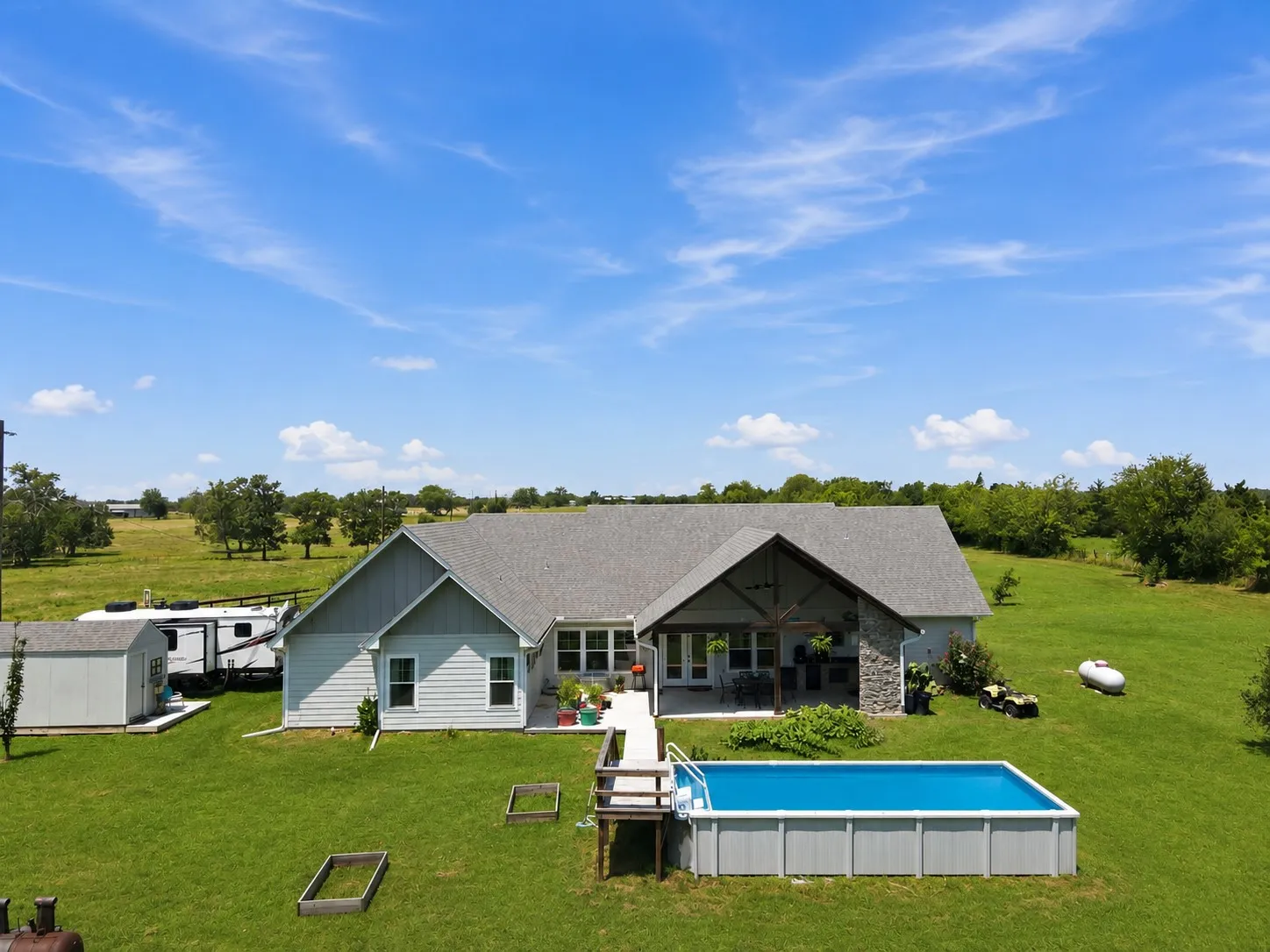 Spacious rear exterior featuring a covered patio with stone accents, an above-ground pool, a detached storage shed, and expansive green lawns