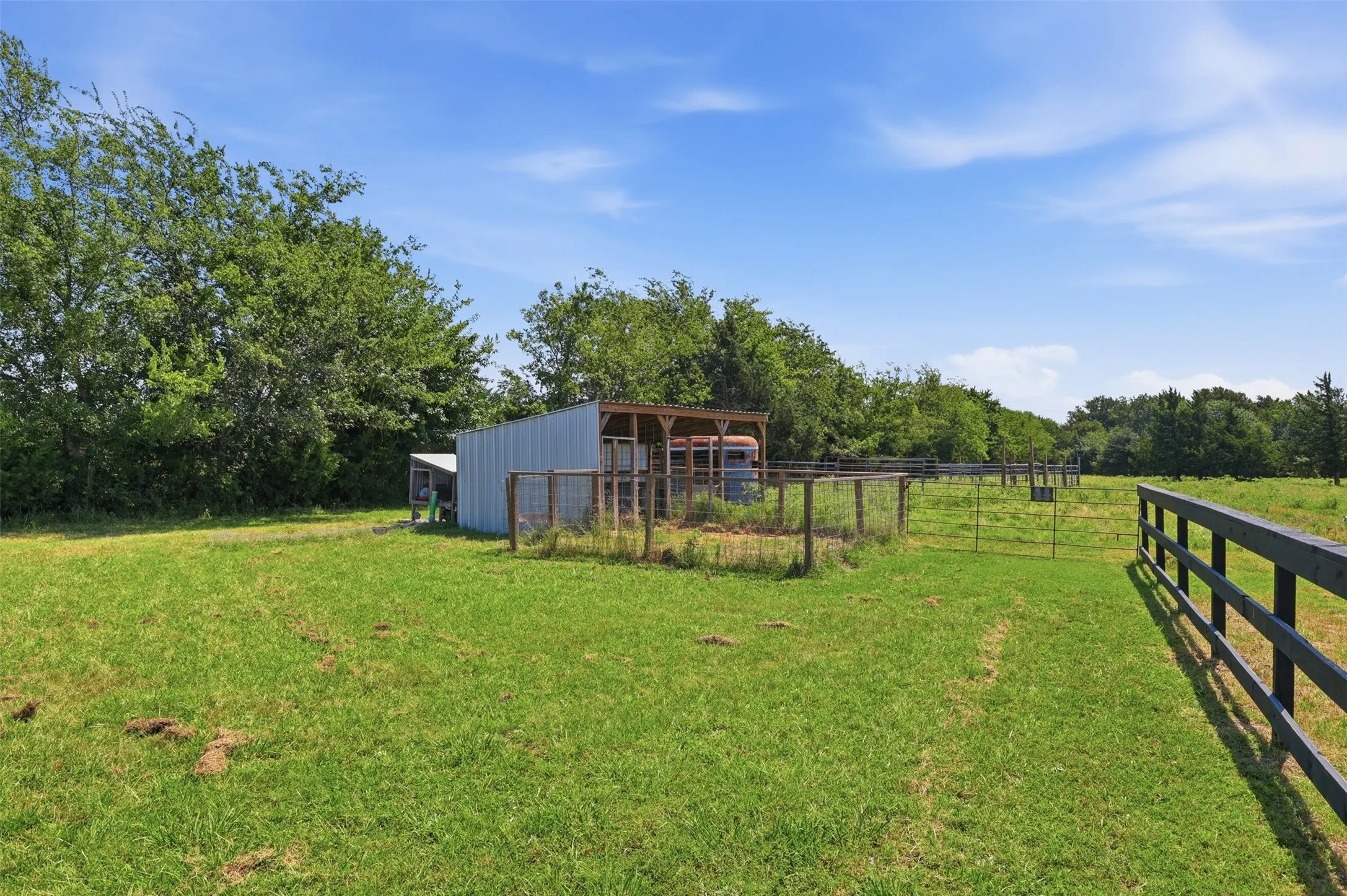 Pastureland featuring a metal outbuilding with a covered section, partial perimeter fencing, and a dark-stained wood fence line