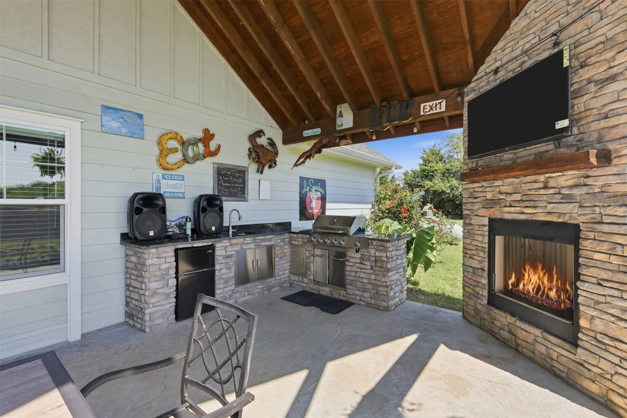Covered outdoor living space featuring an integrated kitchen with a built-in grill and sink, stone-finish countertops, and a beverage refrigerator