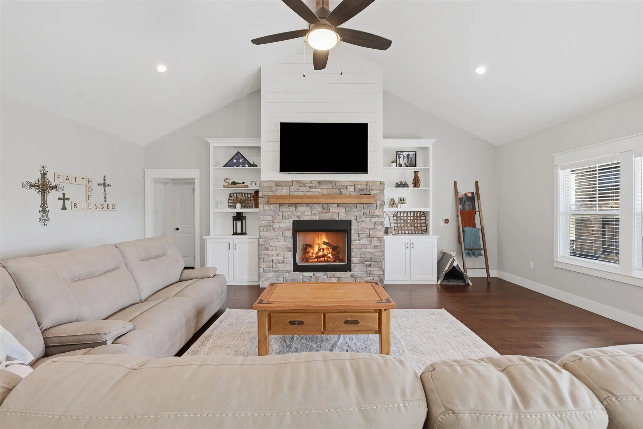 Vaulted living space featuring a stone fireplace with a wood mantel, built-in shelving, wood-finish flooring, and a ceiling fan