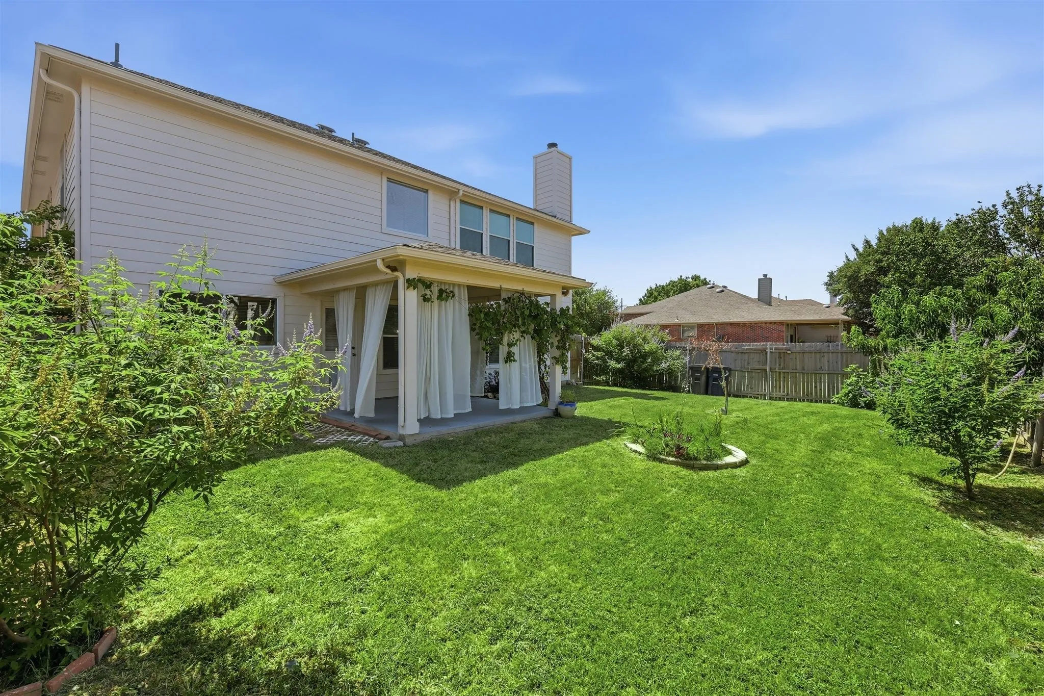 Expansive backyard featuring a large grassy area, mature shrubbery, and a covered patio with privacy curtains