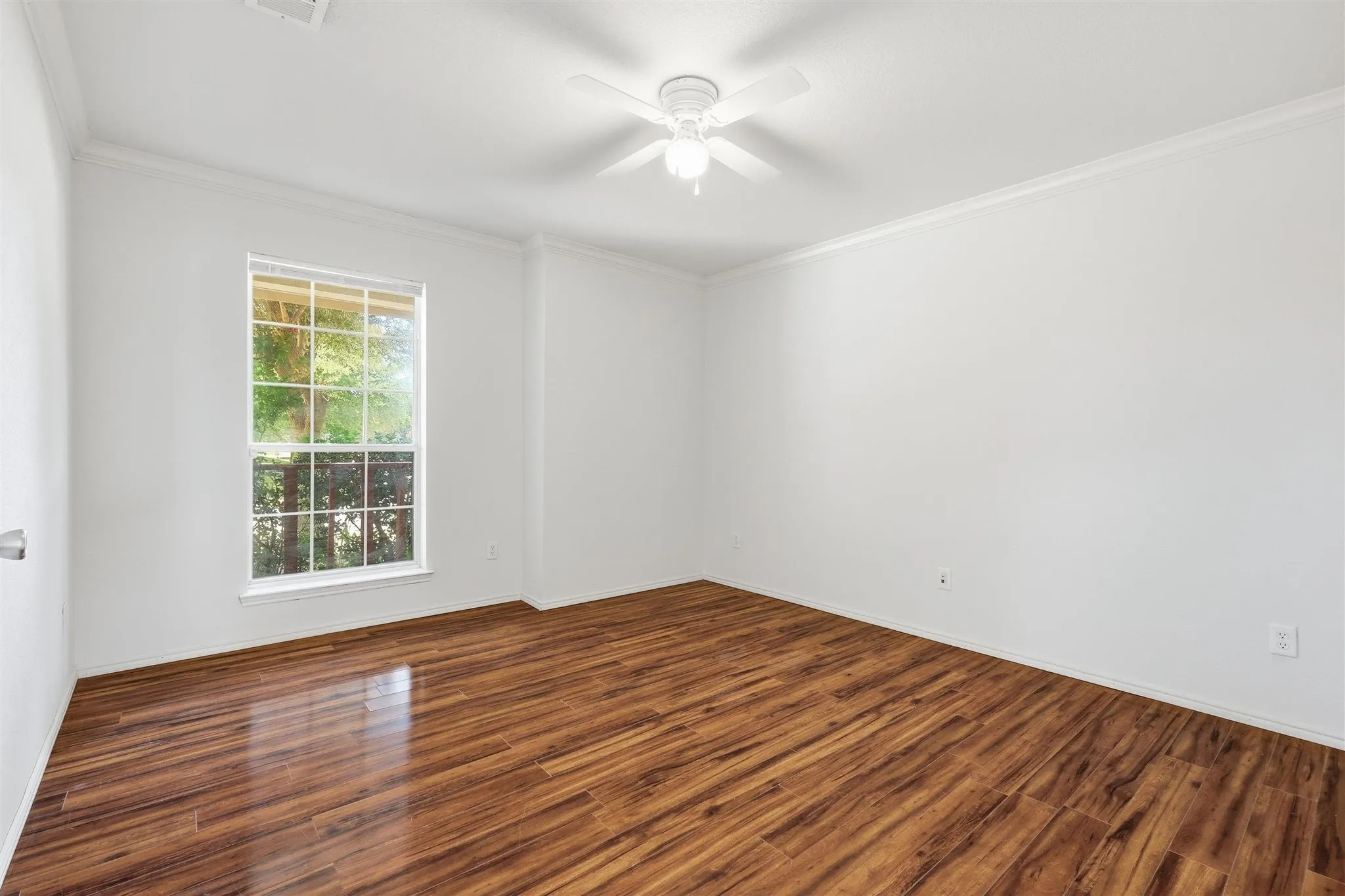 Spacious room featuring wood-finish flooring, a white ceiling fan, crown molding, and a large window with white trim