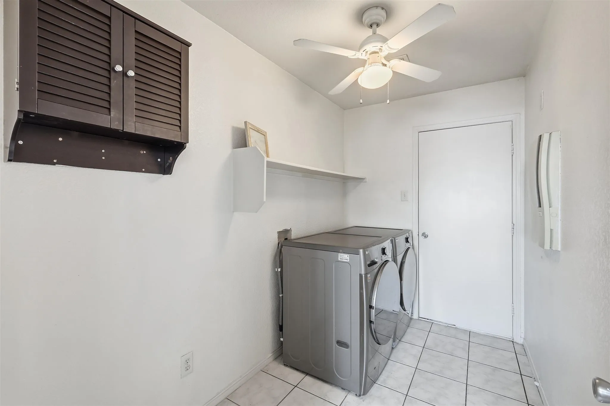 Dedicated laundry area featuring tile flooring, a ceiling fan with integrated lighting, a dark wood louvered cabinet, and a wall-mounted shelving unit