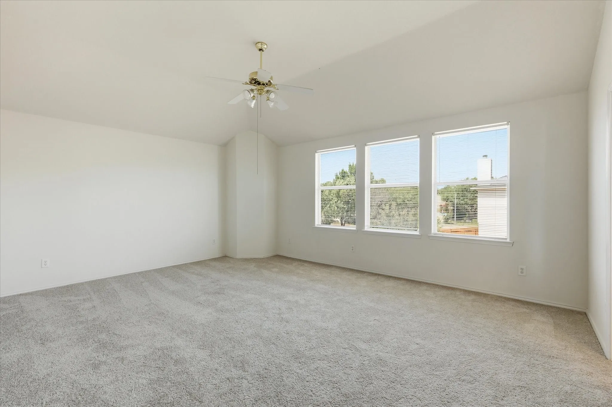 Spacious room featuring vaulted ceilings, a ceiling fan, three large windows with horizontal blinds, and neutral grey carpeting