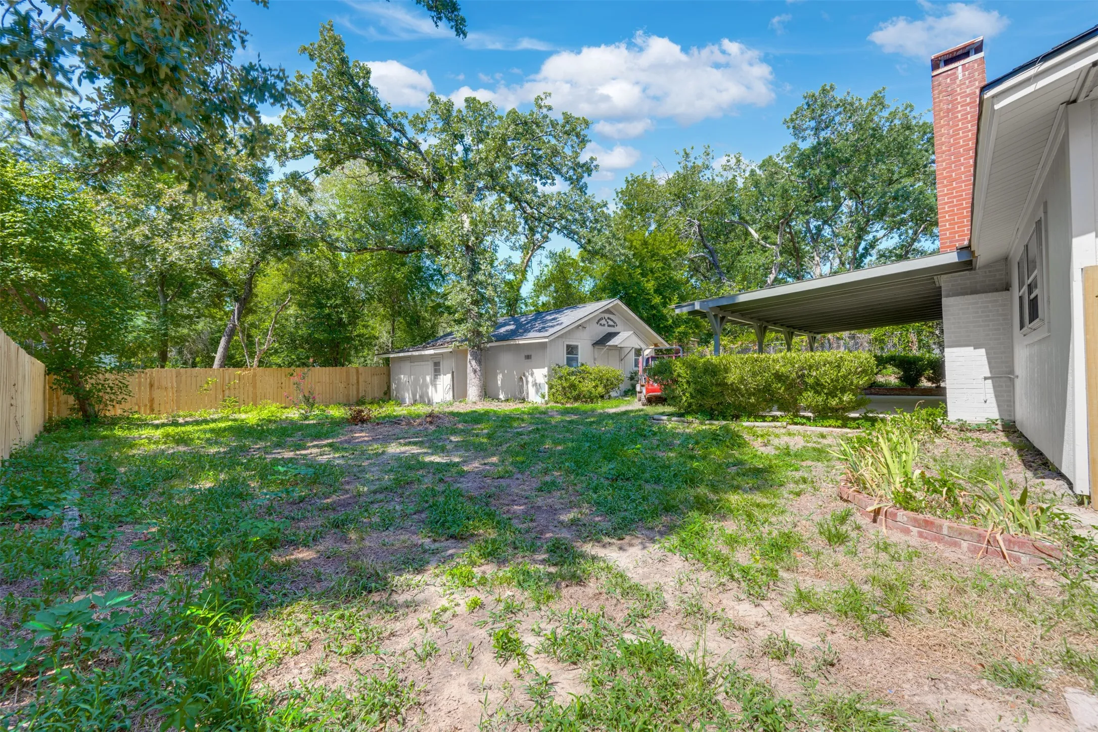 Spacious backyard featuring a new wooden privacy fence, mature trees, a detached auxiliary building, and a covered carport with a brick chimney