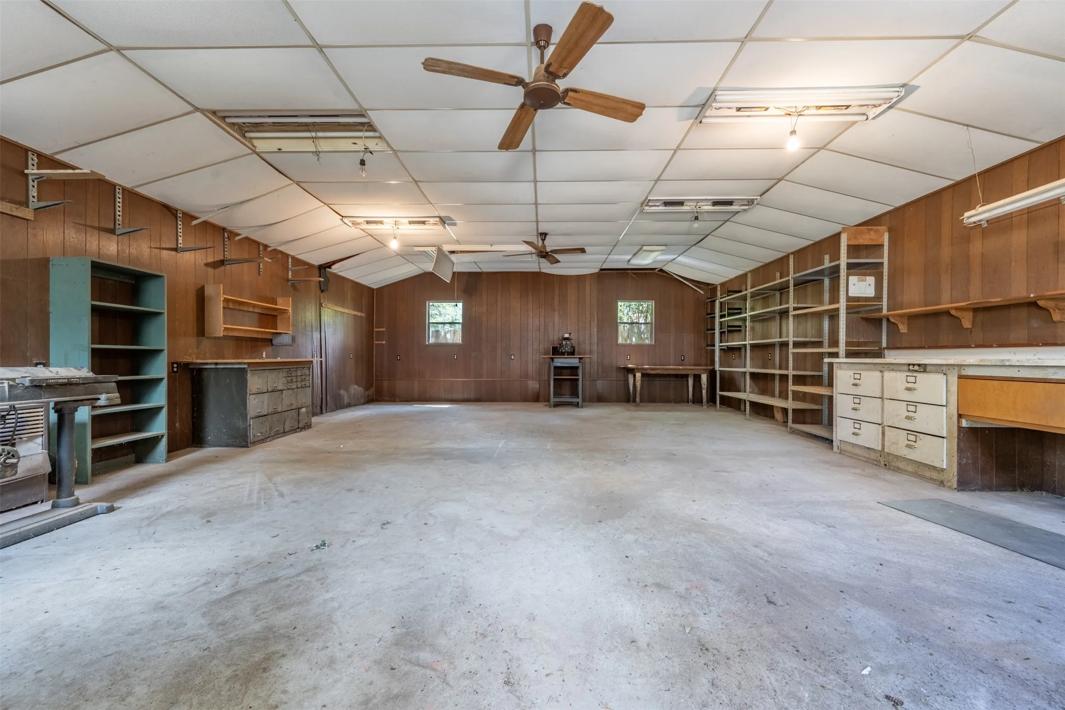Spacious workshop featuring wood-paneled walls, concrete flooring, built-in shelving, and multiple ceiling fans