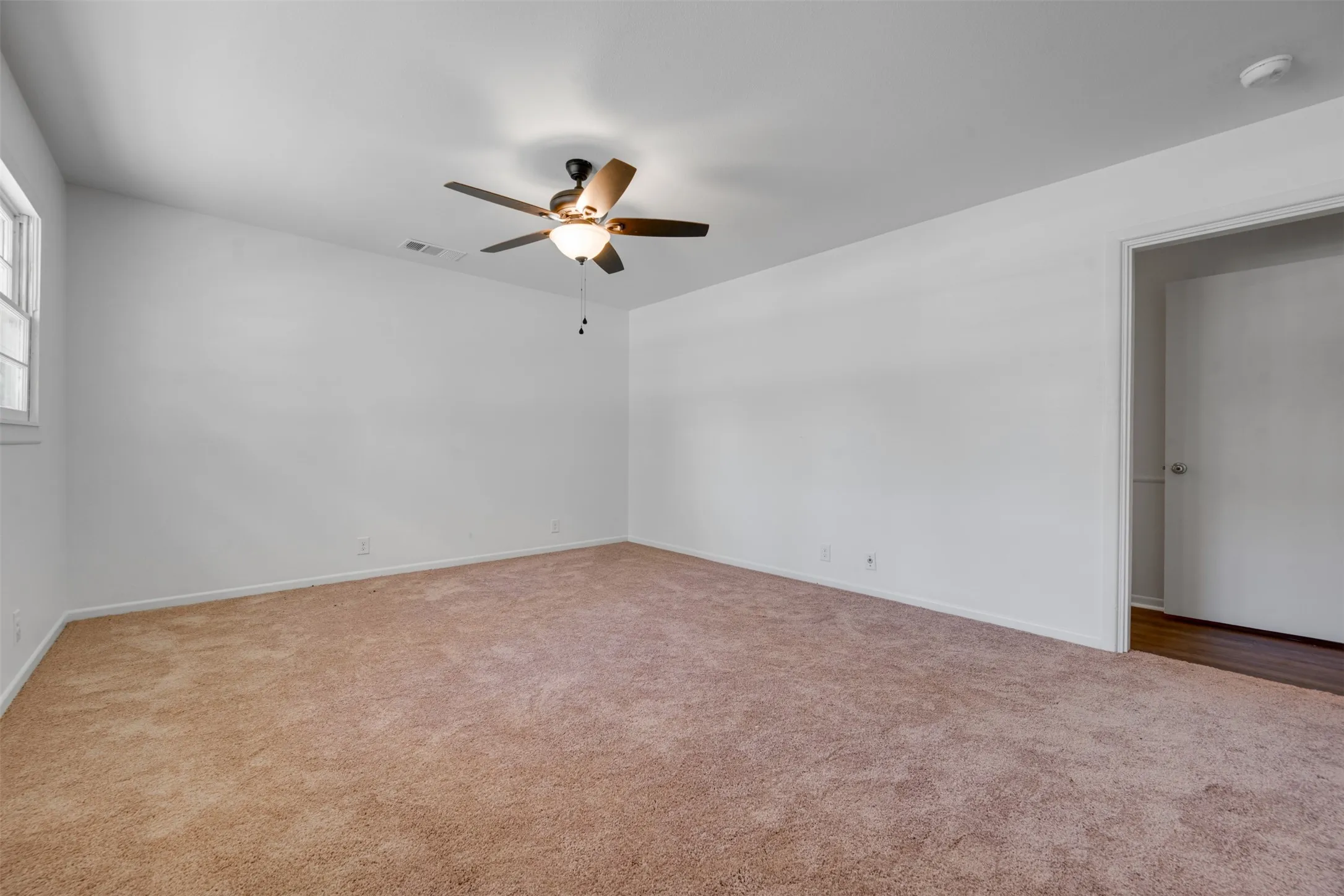 Spacious room featuring neutral carpeting, a ceiling fan with integrated lighting, a window with white trim, and a doorway opening to wood-finish flooring