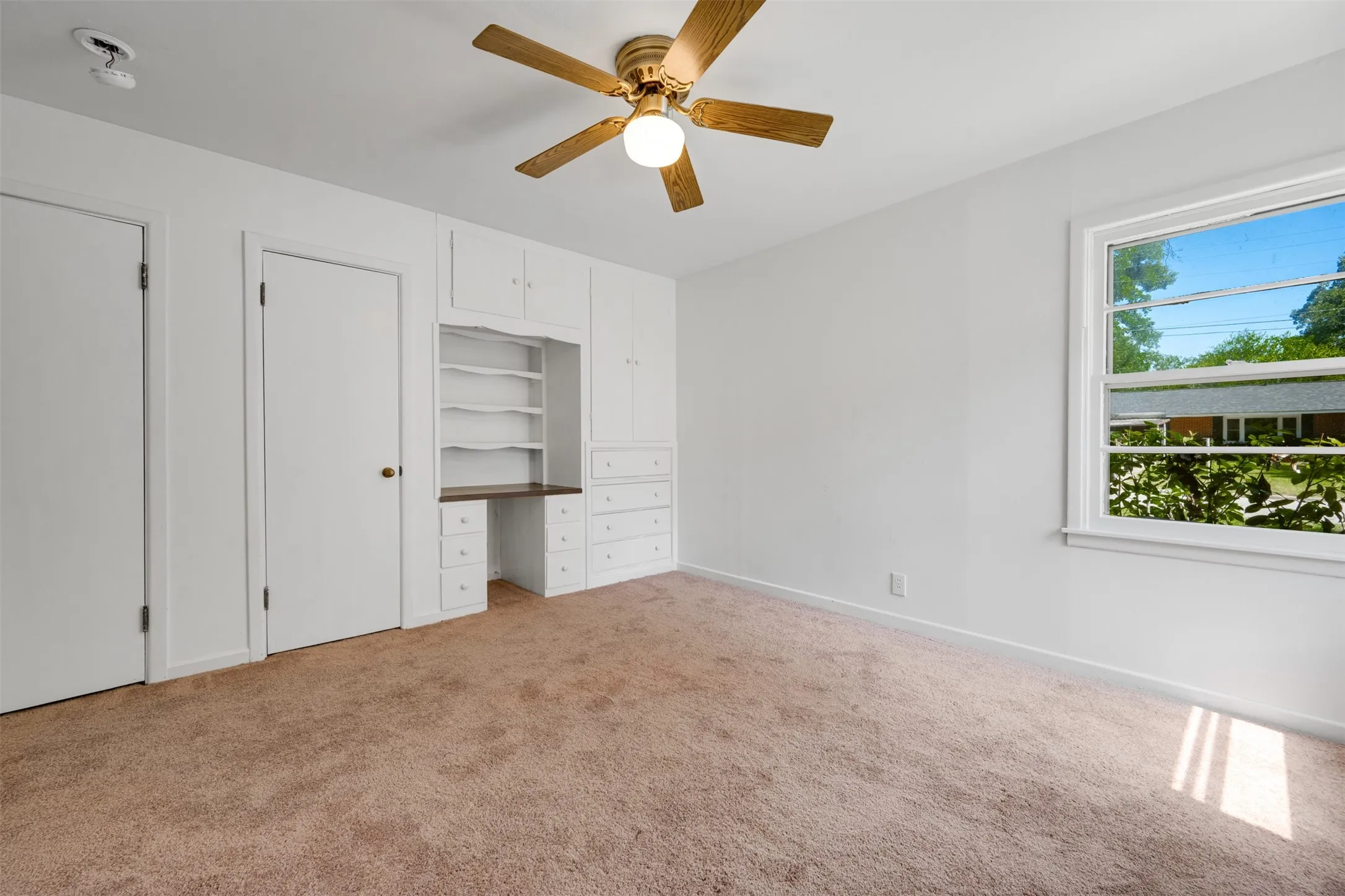 Built-in desk and shelving unit, neutral wall paint, ceiling fan with light fixture, light brown carpet flooring, and a window with exterior views