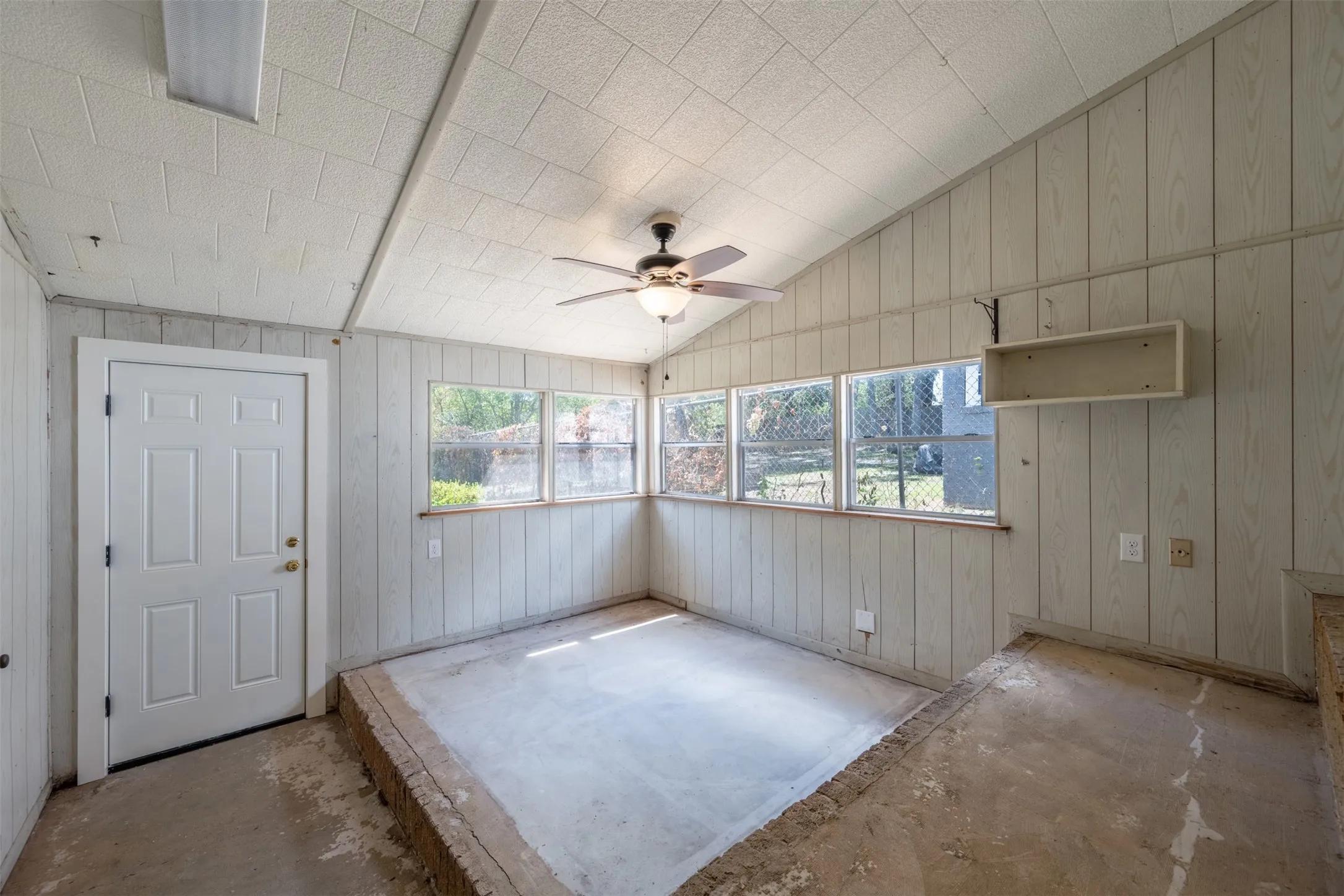 Vaulted ceiling with textured panels, ceiling fan, white paneled walls, multiple windows, and a built-in shelf