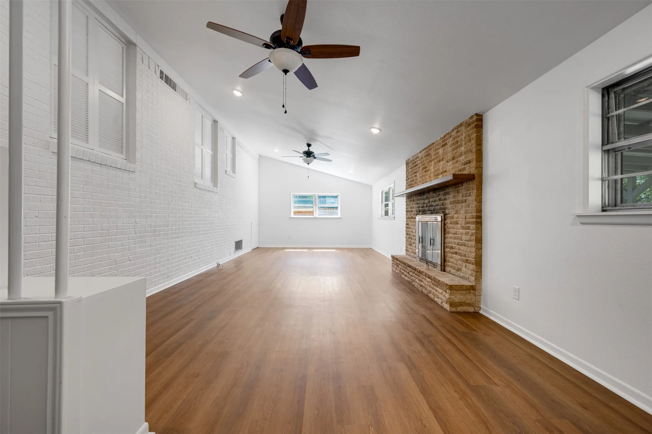 Spacious room with wood-finish flooring, white painted brick accent wall, brick fireplace with metal insert and wood mantle, vaulted ceiling, and dual ceiling fans