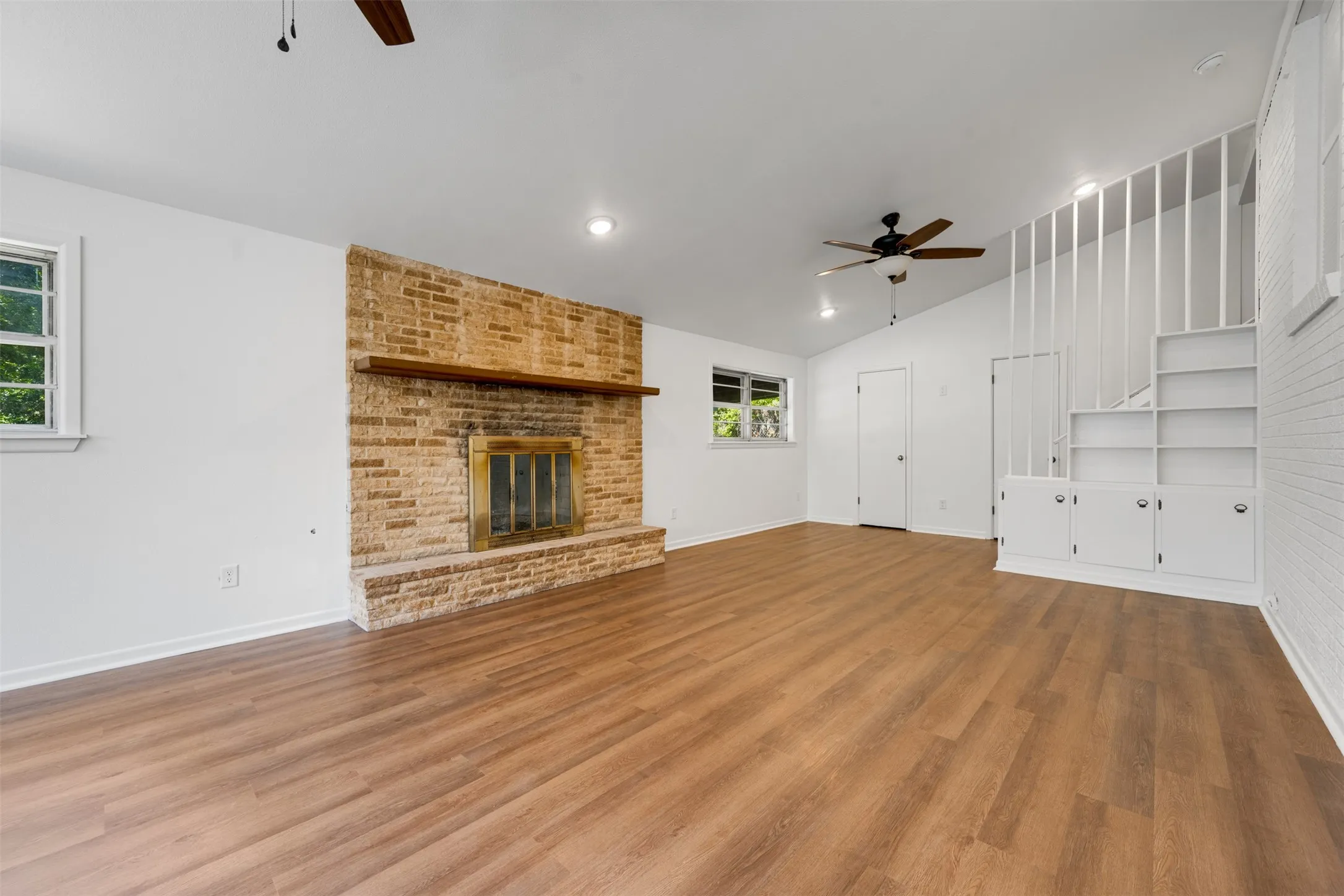 Spacious living area featuring wood-finish flooring, a prominent brick fireplace with a wood mantle, and built-in shelving with cabinetry