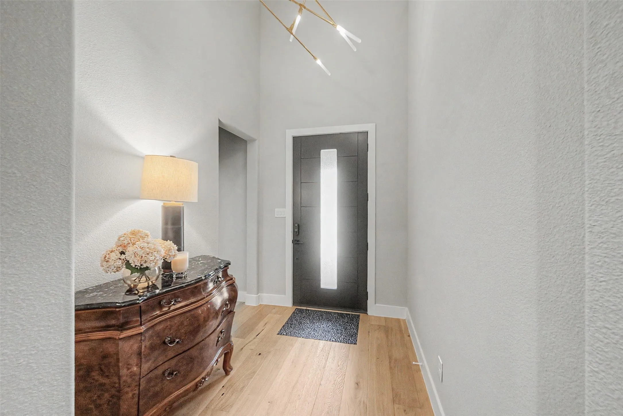 Foyer featuring a dark-paneled door with a vertical frosted glass insert, light wood-finish flooring, a modern linear chandelier, and textured white walls