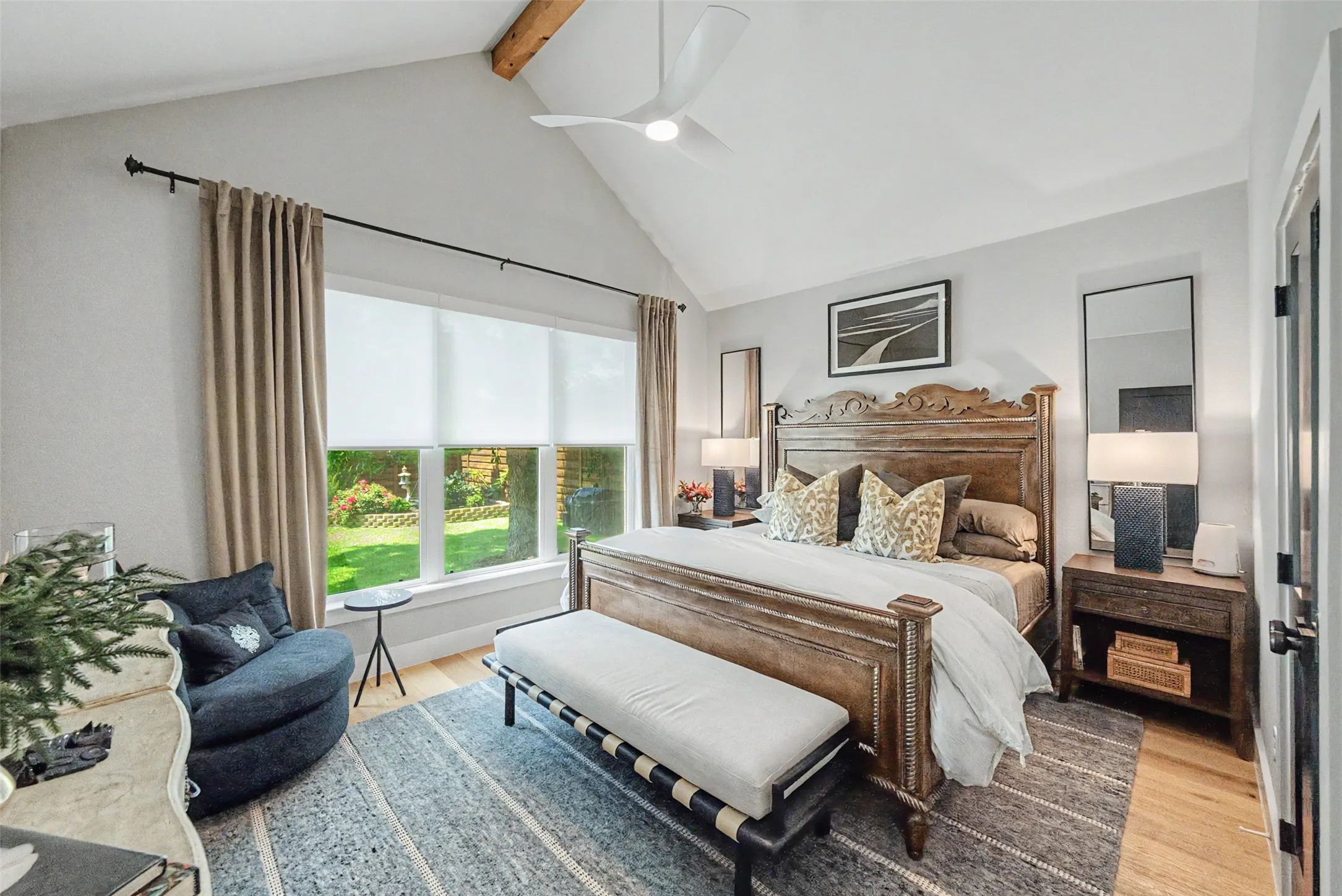 Spacious room featuring a vaulted ceiling with exposed wood beam, a large window overlooking a manicured outdoor area, light wood-finish flooring, and a ceiling fan
