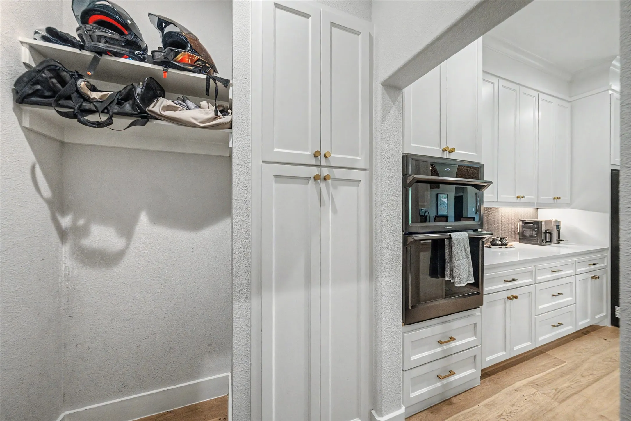 Modern kitchen featuring white shaker cabinetry, brass hardware, built-in double ovens, and light-toned wood-finish flooring
