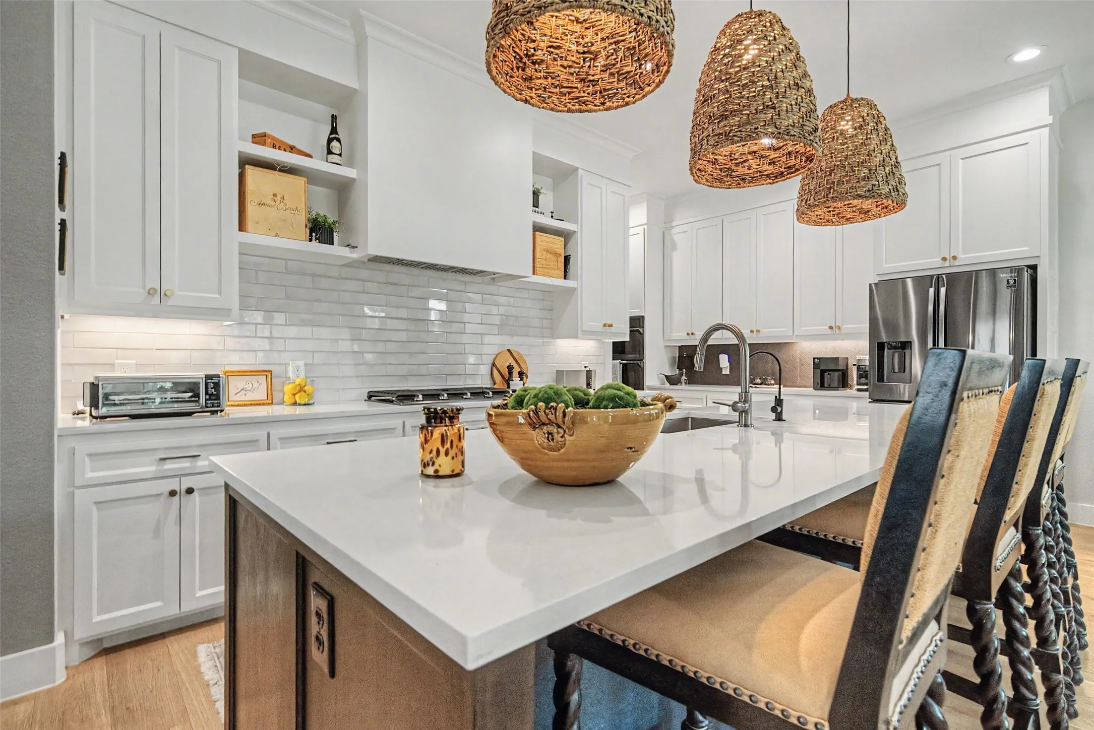 Modern kitchen featuring a large island with a light-toned countertop, white shaker-style cabinetry, a subway tile backsplash, stainless steel appliances, and wood-finish flooring