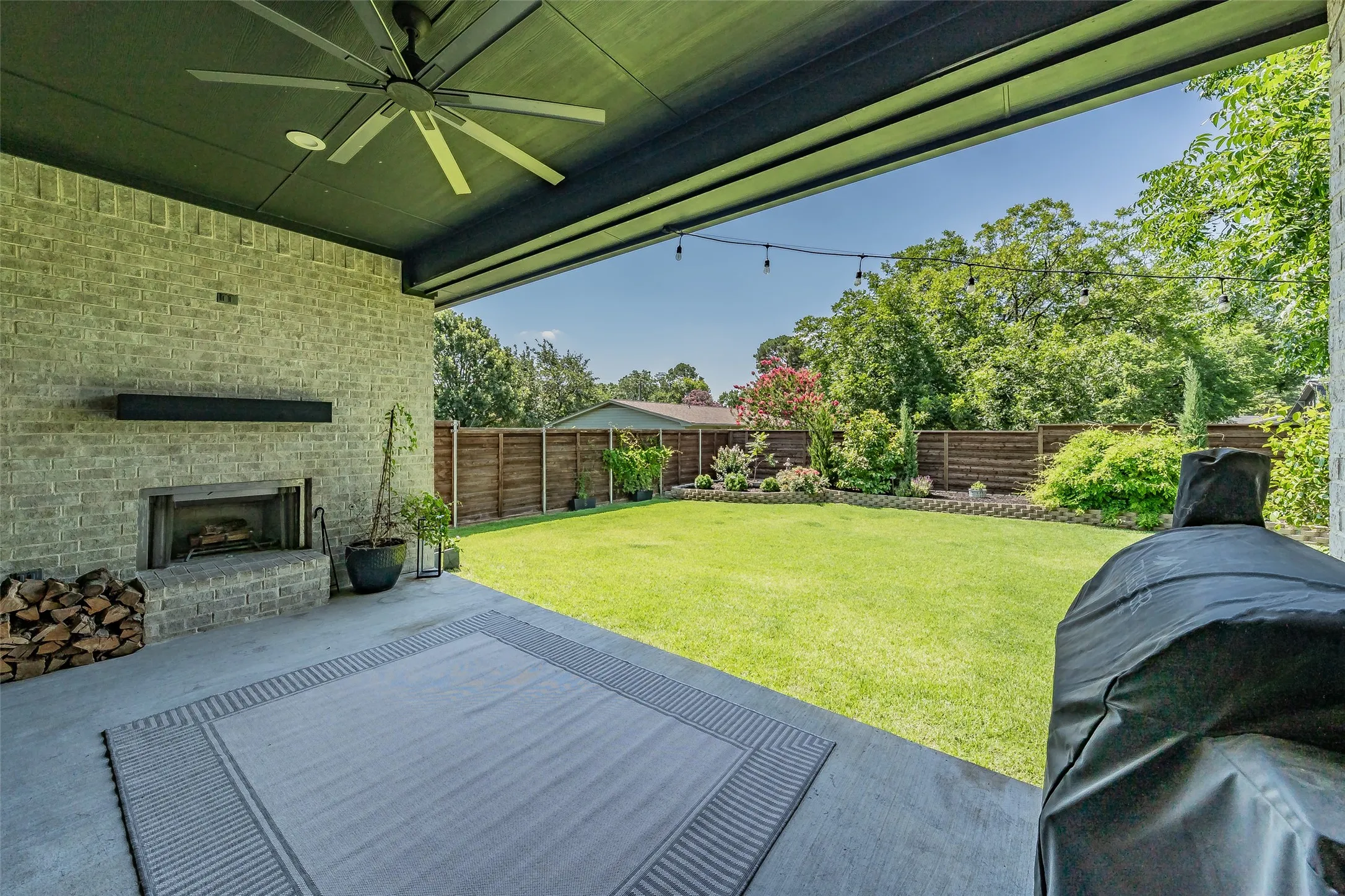 Covered outdoor patio featuring a brick fireplace, built-in ceiling fan, string lighting, and a spacious grassy yard
