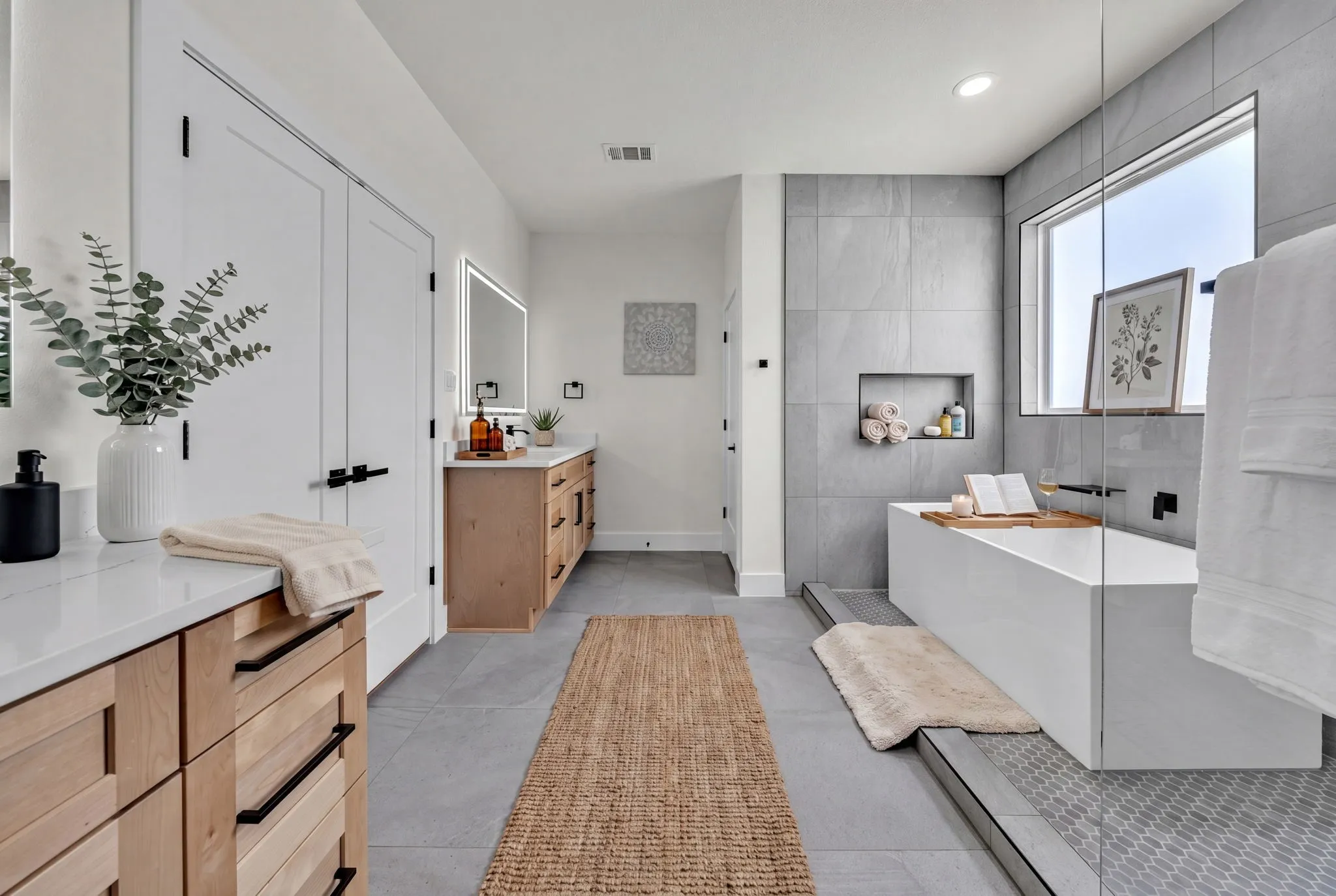 Contemporary bathroom featuring a freestanding soaking tub, a dual vanity with light wood cabinetry, a walk-in shower with a glass enclosure, large format gray wall tiling, and a built-in shower niche