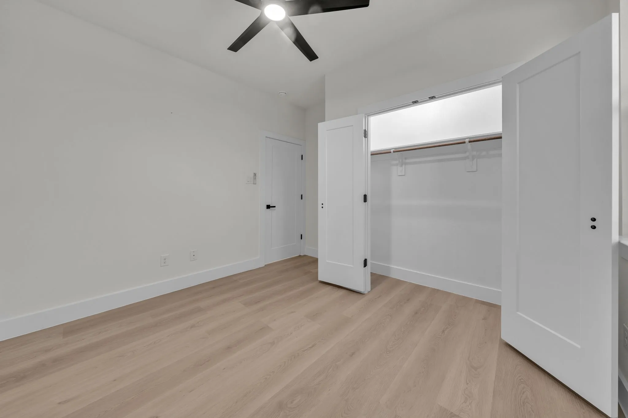 Bedroom featuring light wood-finish flooring, a contemporary ceiling fan with black blades, white baseboards, and a spacious closet with double doors and a built-in hanging rod