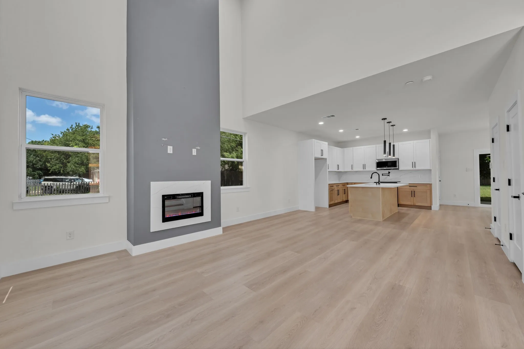 Open-concept living space featuring wood-finish flooring, a contemporary fireplace with a contrasting accent wall, and a kitchen with white upper cabinetry, wood-finish lower cabinetry, and a central island with a sink and pendant lighting