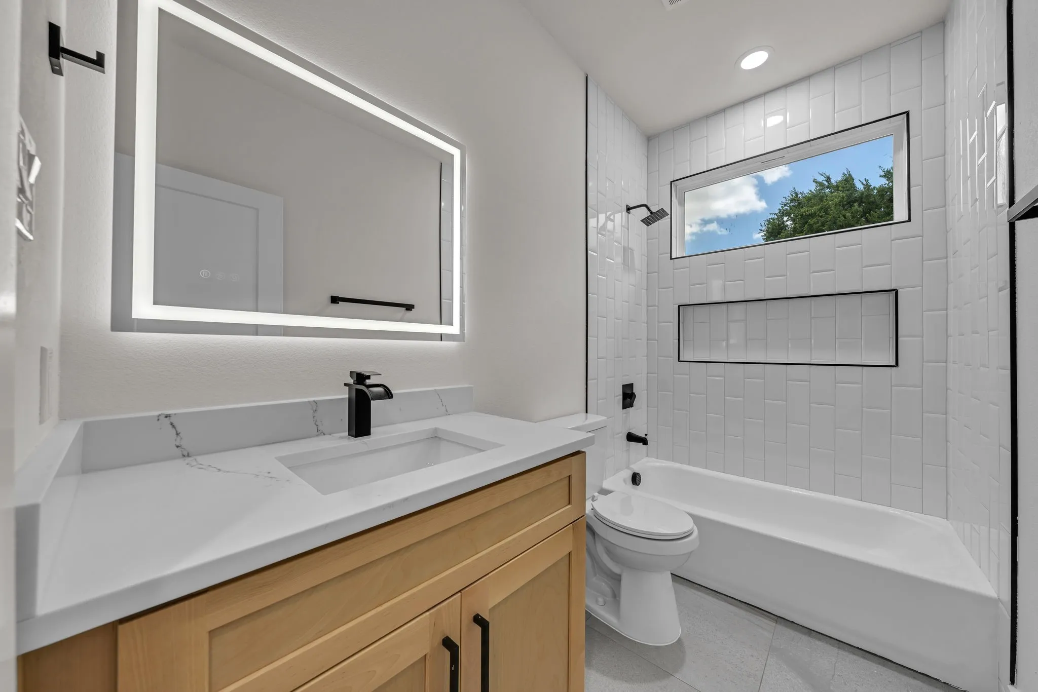 Contemporary bathroom featuring a light wood-finish vanity with a white countertop and integrated sink, illuminated by a rectangular backlit mirror