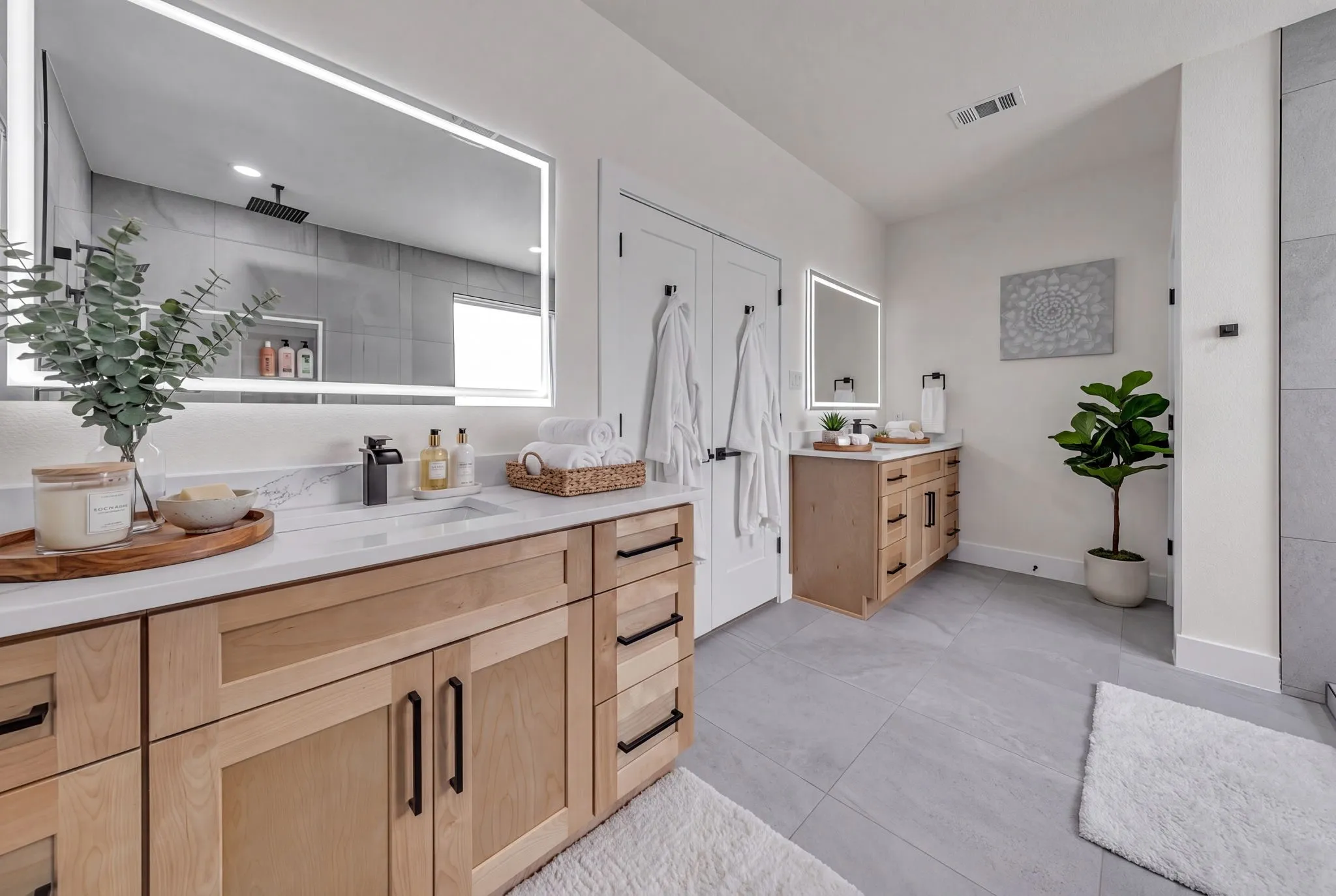 Double vanity bathroom featuring light wood cabinetry with black hardware, white countertops, illuminated mirrors, and light gray tile flooring