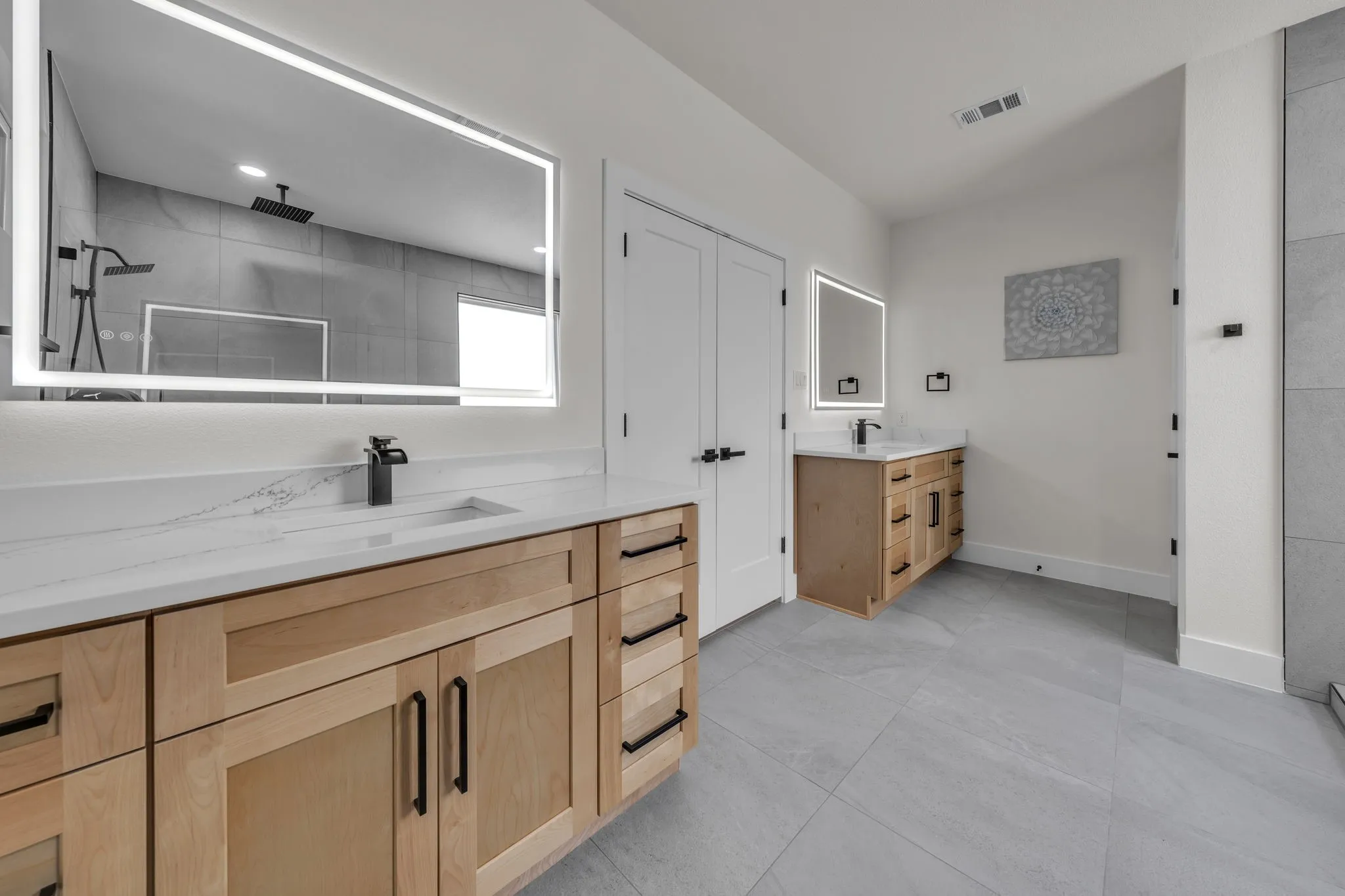 Spacious bathroom featuring light wood cabinetry with matte black hardware, white countertops, illuminated mirrors, and large format grey floor tiling