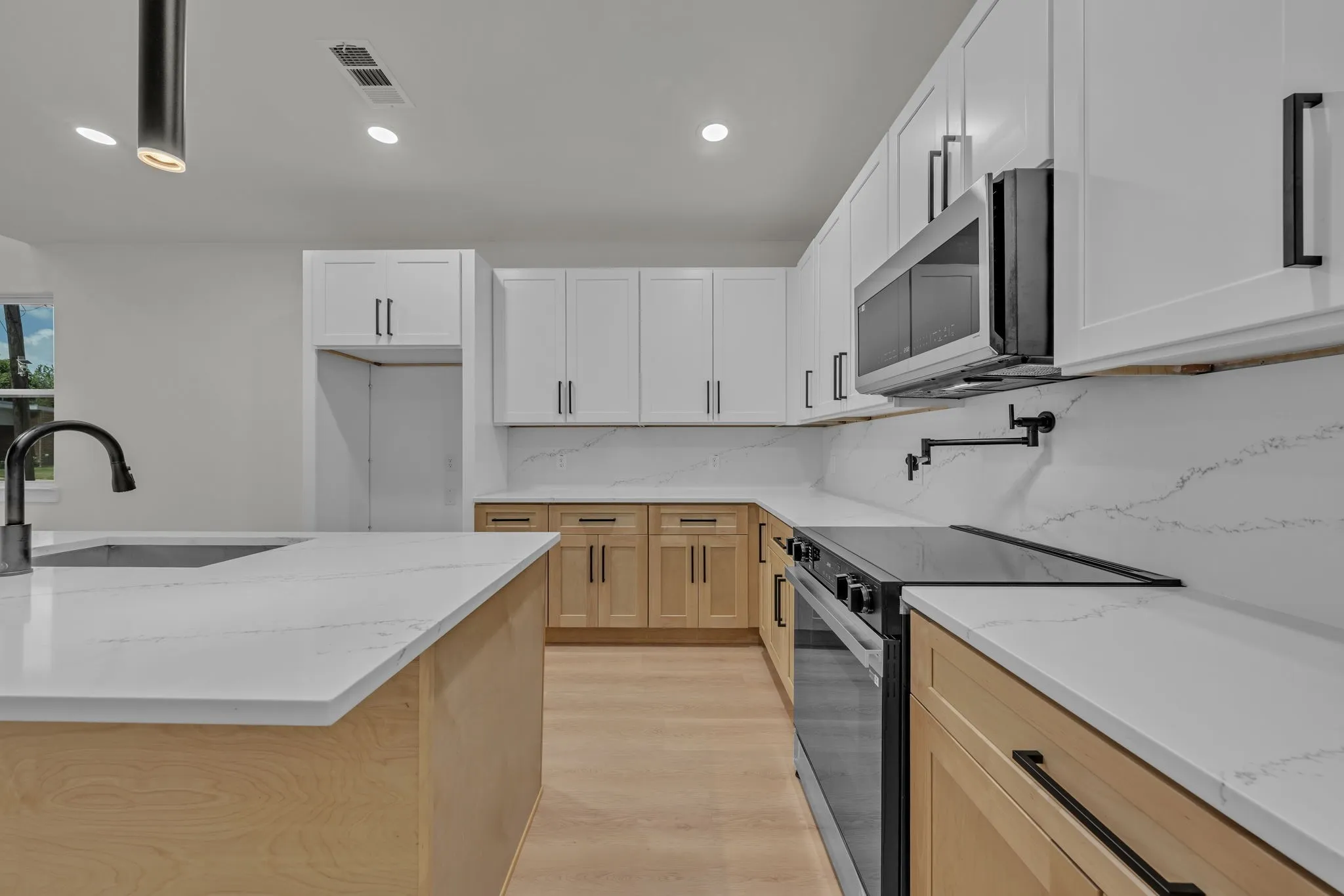Kitchen featuring a central island with an undermount sink and matte black faucet, light wood-finish lower cabinetry, white upper cabinetry, and white countertops with a patterned backsplash