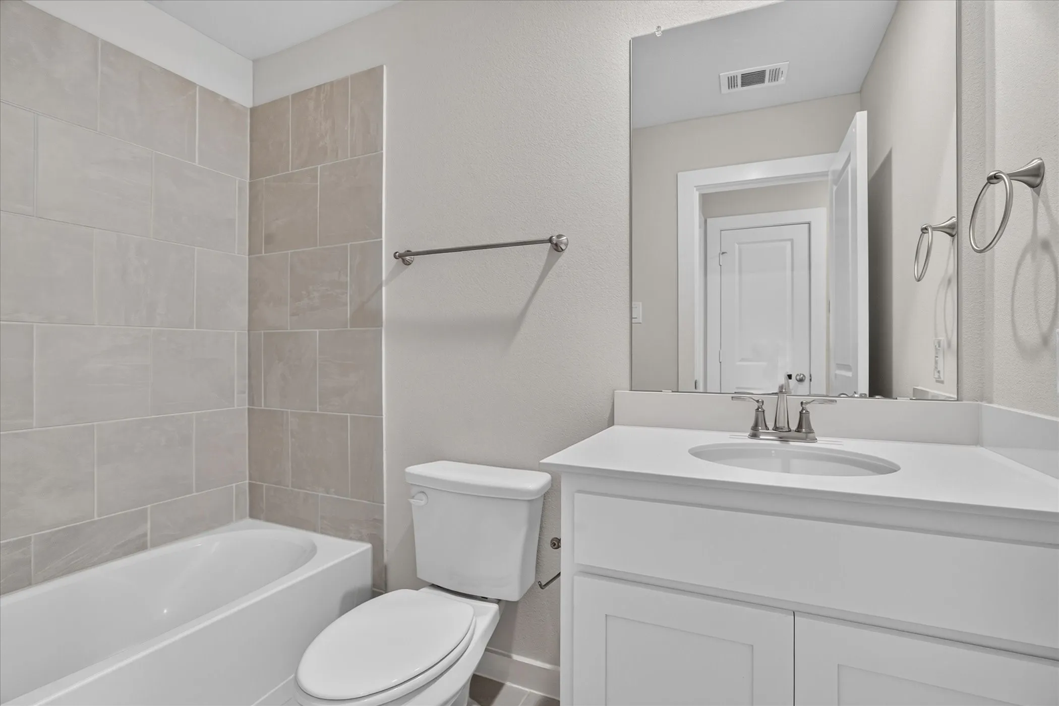 White vanity with integrated sink and chrome faucet, large frameless mirror, white bathtub with light-toned tiled surround, and white toilet