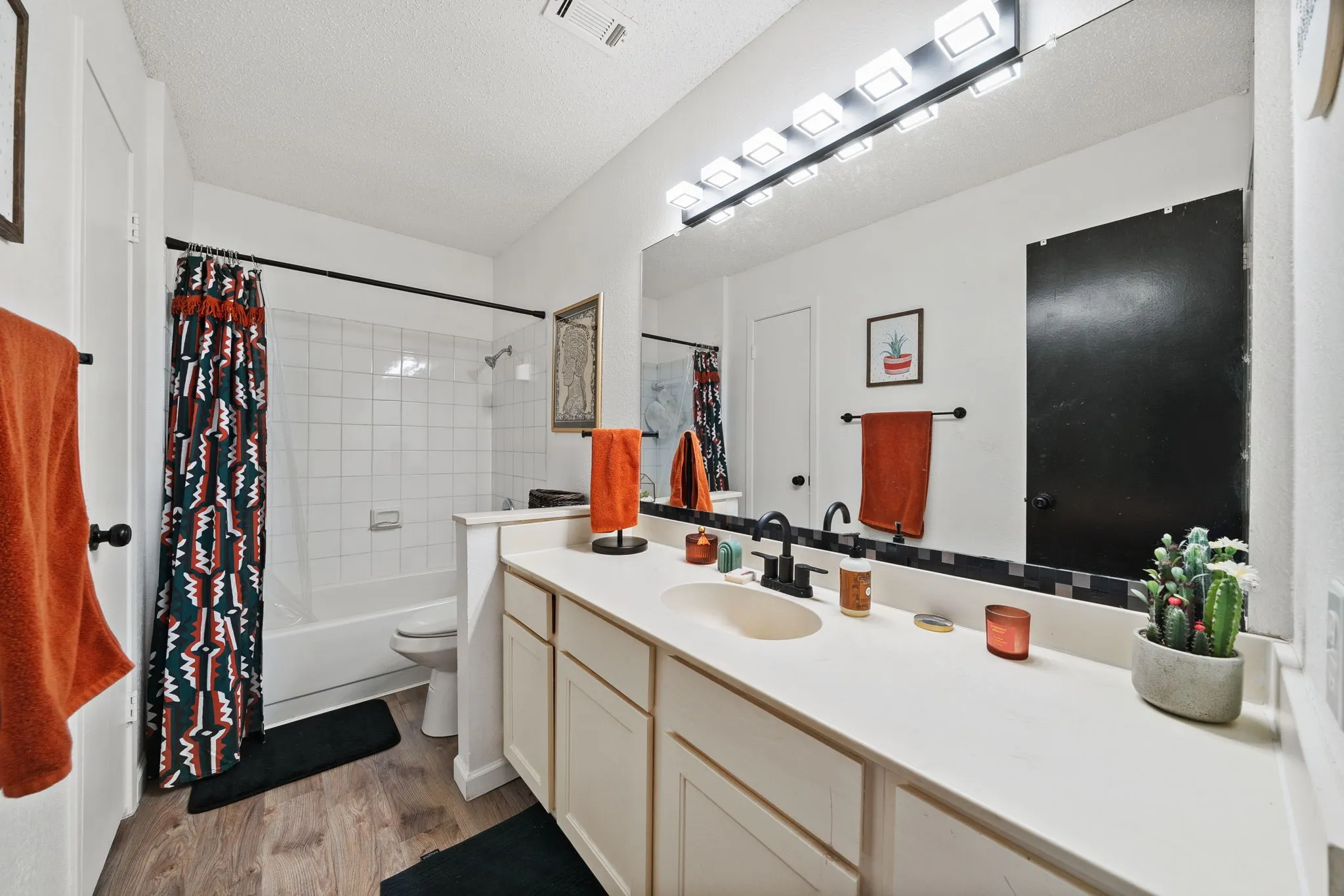 Bathroom featuring wood-finish flooring, a white vanity with dual sinks, a large mirror, a bath/shower combination with white subway tile surround, and black fixtures