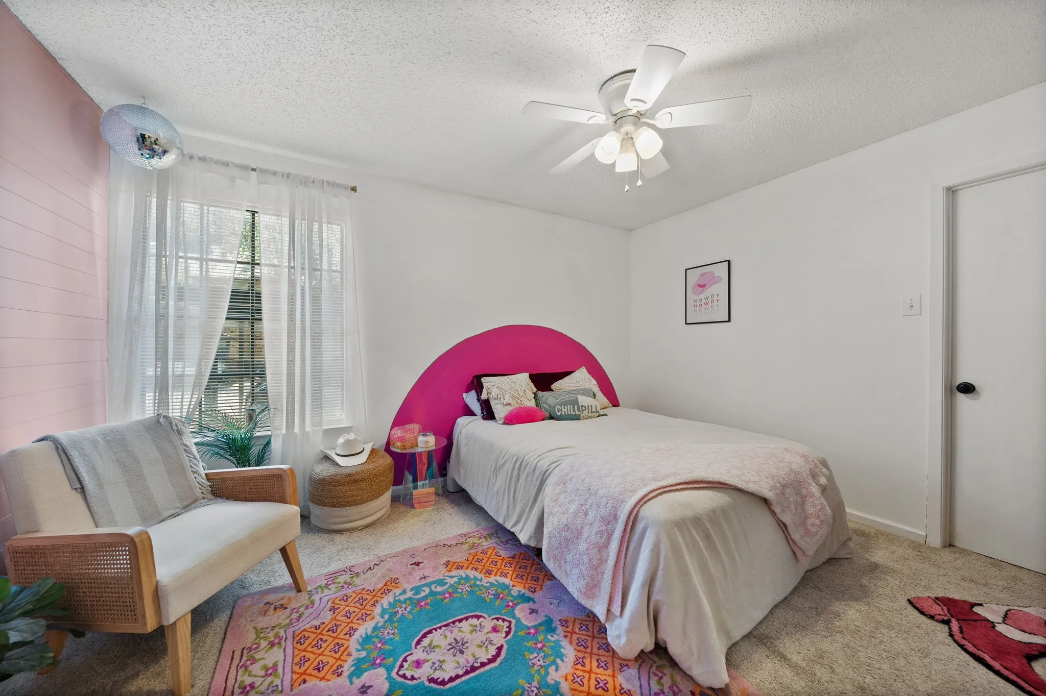 Bedroom featuring a window with natural light, textured pink accent wall, white ceiling fan with light kit, and neutral carpeting