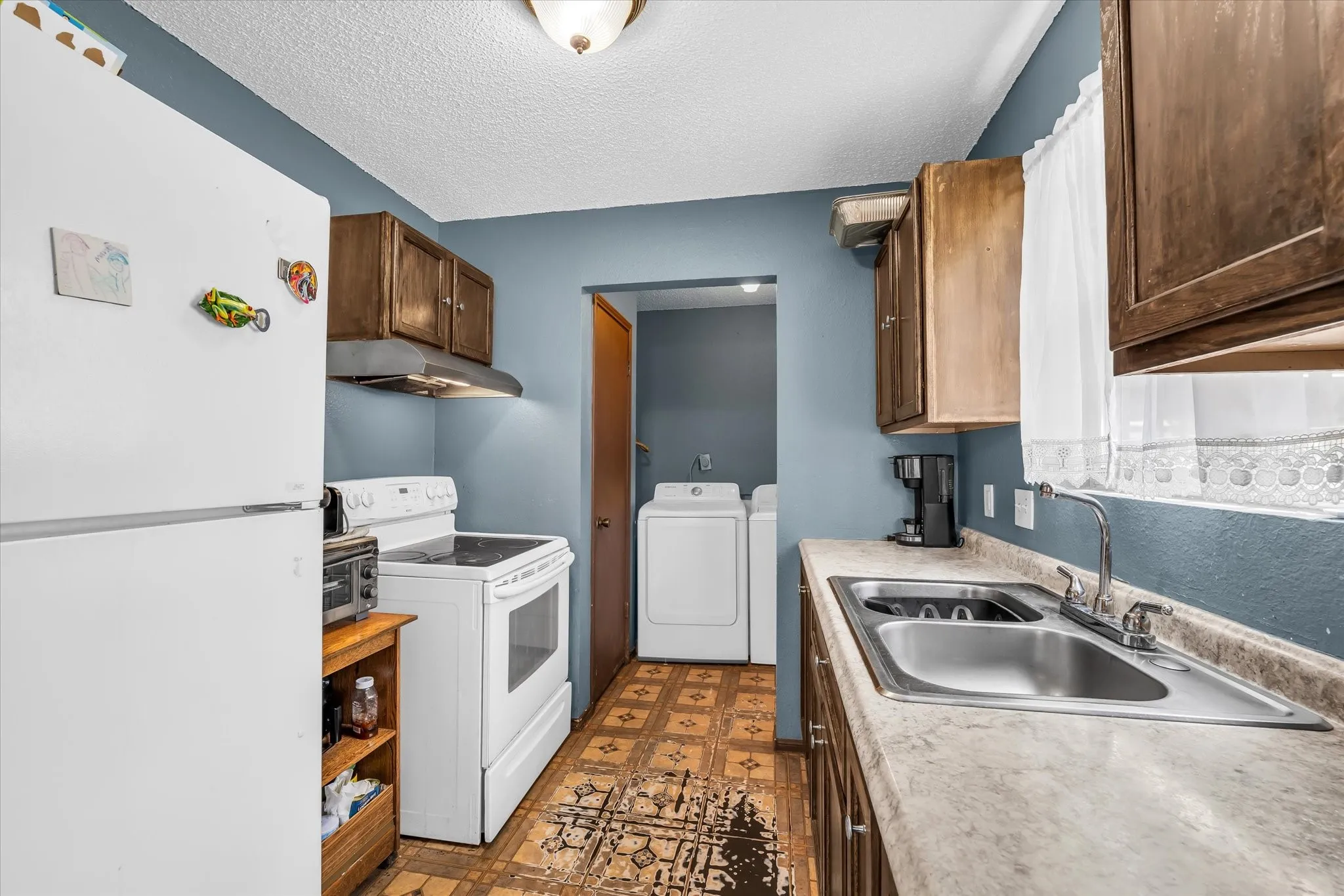 Kitchen area featuring white appliances, wood cabinetry, a double basin stainless steel sink, and a light-toned countertop