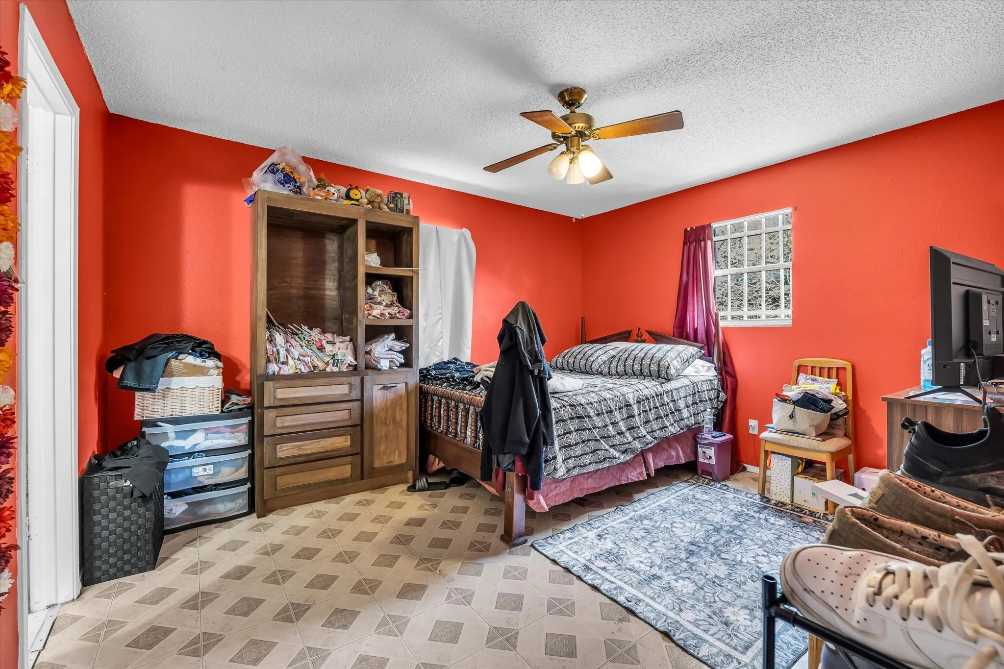 Bedroom featuring red walls, a ceiling fan with light fixture, a window with vertical bars, and patterned tile-style flooring