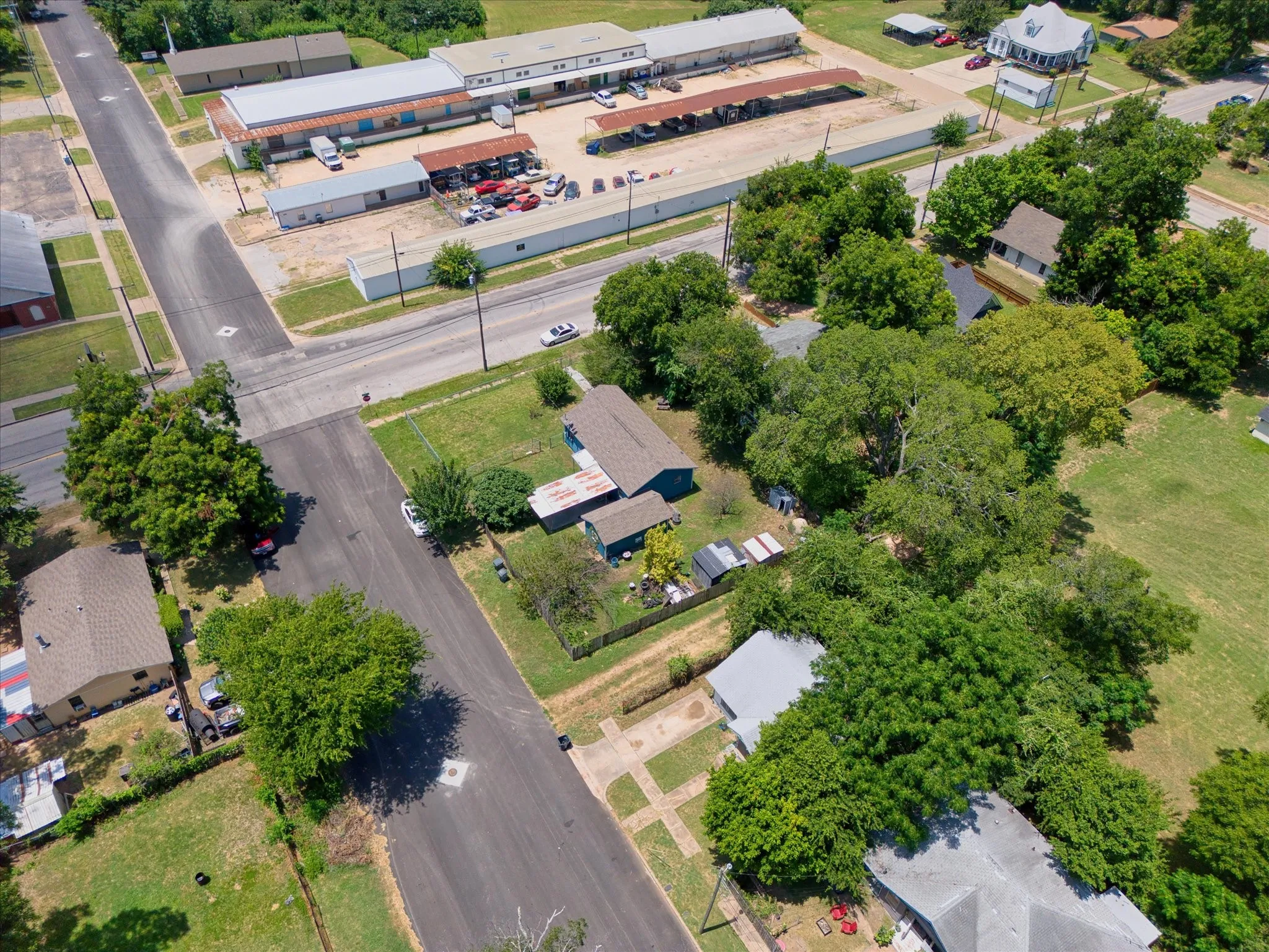 Aerial view showcasing a residential property with surrounding mature trees, a paved street, and a nearby commercial area