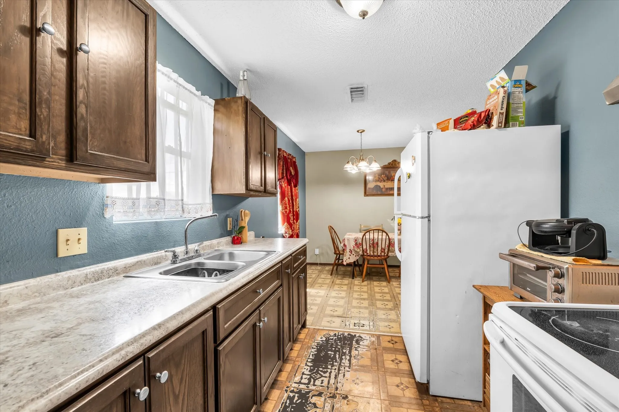 Galley-style kitchen featuring wood cabinetry, a double basin stainless steel sink, light-toned countertops, and an integrated white refrigerator