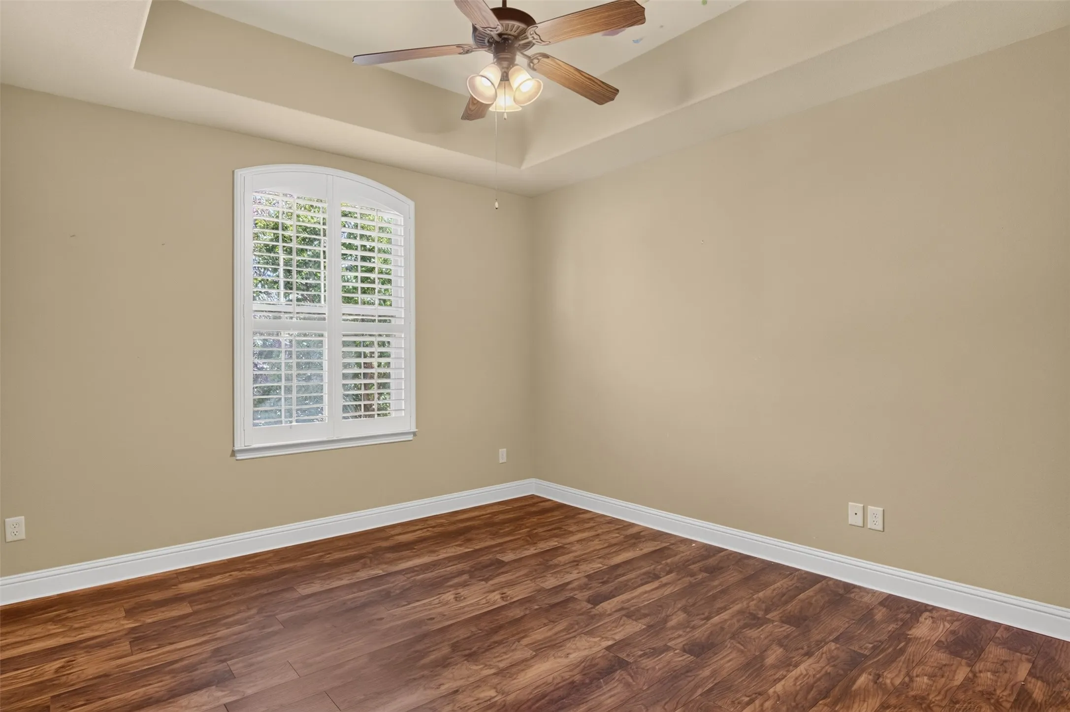 Bedroom featuring tray ceiling with a ceiling fan, wood-finish flooring, and arched window with plantation shutters