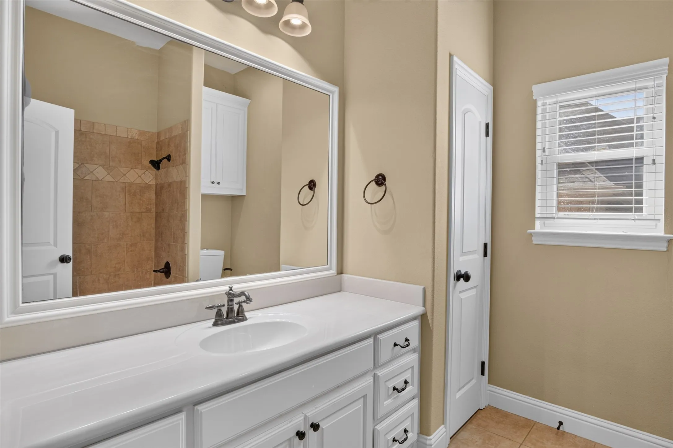 Bathroom featuring a vanity with a white countertop and integrated sink, cabinet storage, a large mirror, and a window with blinds