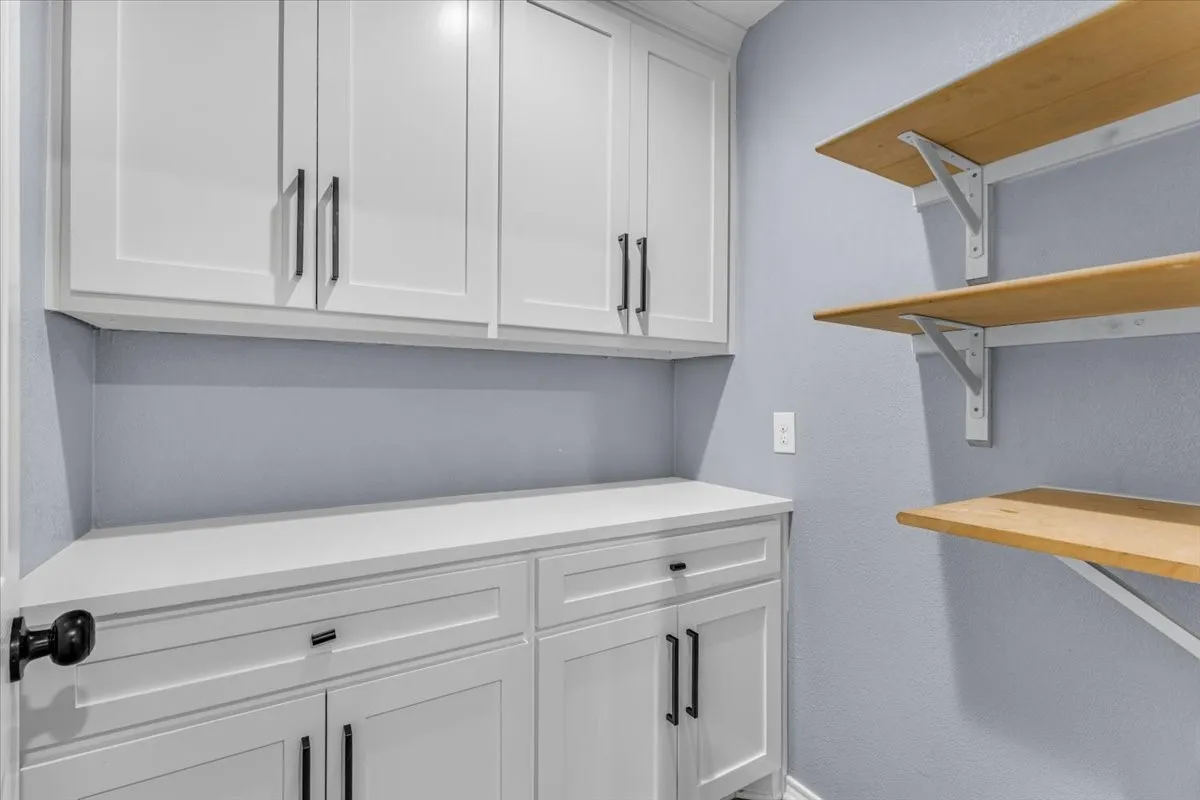 This highly functional utility or pantry space offers excellent organization, featuring crisp white cabinetry with sleek black hardware, ample counter space, and sturdy, open shelving.