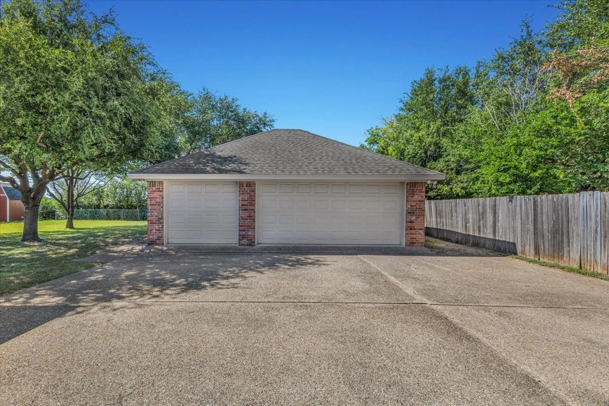 A substantial detached three-car brick garage sits at the end of an expansive concrete driveway, providing tons of storage space and ample room for a large SUV or extended truck. The structure features a single and a double bay door, perfectly complementing the property's mature landscaping and solid wooden privacy fence.