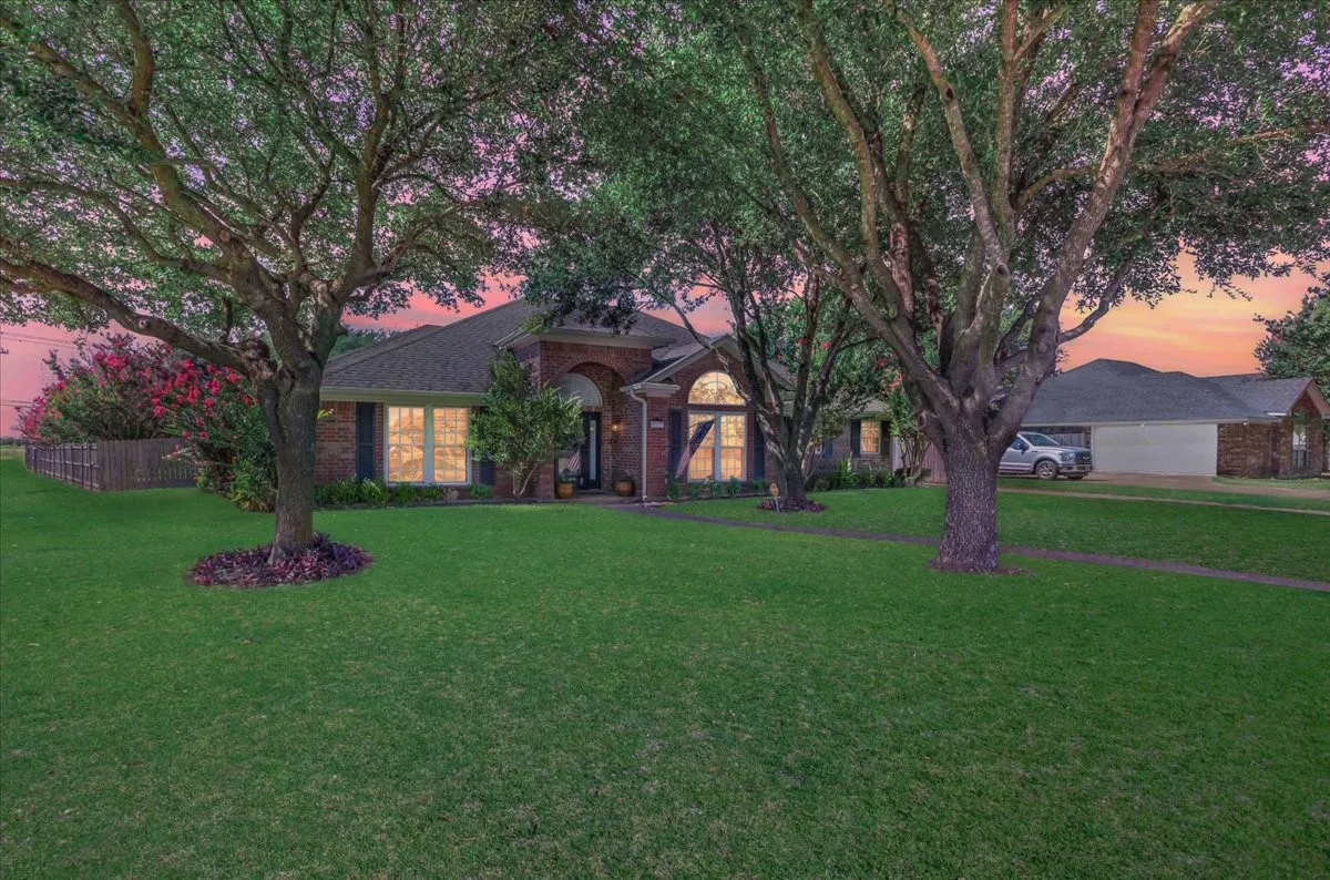 Captured at twilight, this stunning brick home exudes immense curb appeal, perfectly framed by majestic mature trees and a sprawling, manicured lawn. The warm glow from the prominent arched windows creates an inviting ambiance against the vibrant evening sky, beautifully highlighting the property's classic architecture, vibrant blooming crepe myrtles, and the long driveway leading to the detached garage.