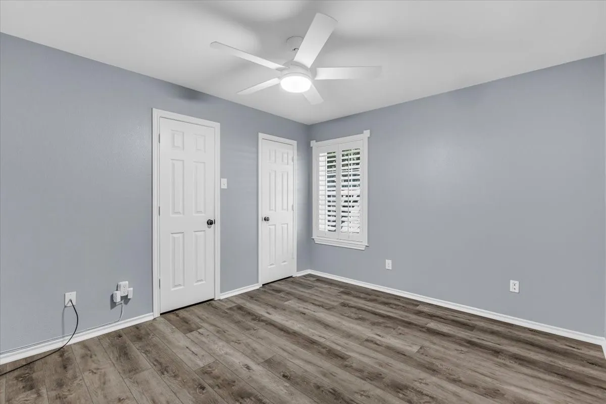A bright and inviting third bedroom featuring crisp neutral paint, durable luxury vinyl plank flooring, a modern lighted ceiling fan, and classic plantation shutters.
