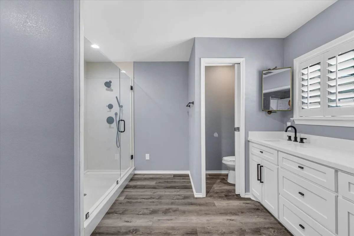 This luxurious first primary bathroom offers a spa-like retreat, featuring a sleek white vanity with modern matte black hardware.  The highlight of the space is a stunning oversized frameless glass walk-in shower equipped with a premium multi-head shower system, all beautifully complemented by continuous luxury vinyl flooring and custom plantation shutters.