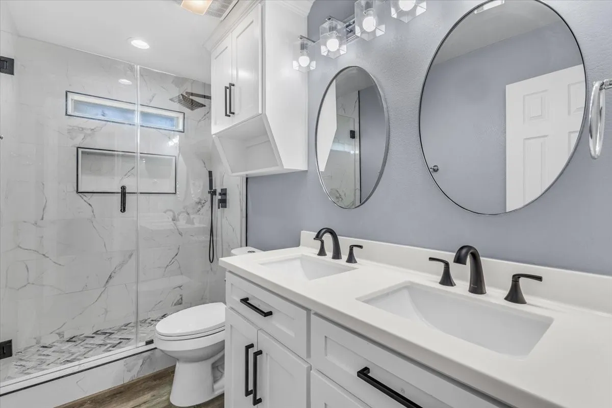 This beautifully renovated bathroom features a sleek double vanity with crisp white cabinetry and striking matte black hardware. The space is anchored by a stunning glass-enclosed walk-in shower, showcasing luxurious marble-look tile, a custom herringbone floor, a built-in niche, and premium black fixtures including a rain showerhead. Dual round mirrors and modern lighting complete this elegant, modern retreat.