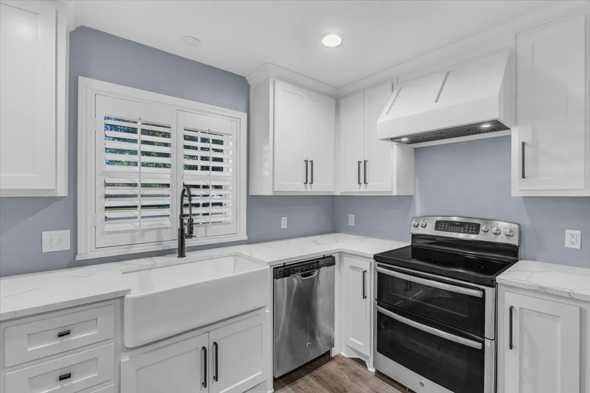 This beautifully updated kitchen features a classic farmhouse sink with a modern black faucet, quartz countertops, a stylish custom range hood, and a stainless steel stove that will remain with the property.