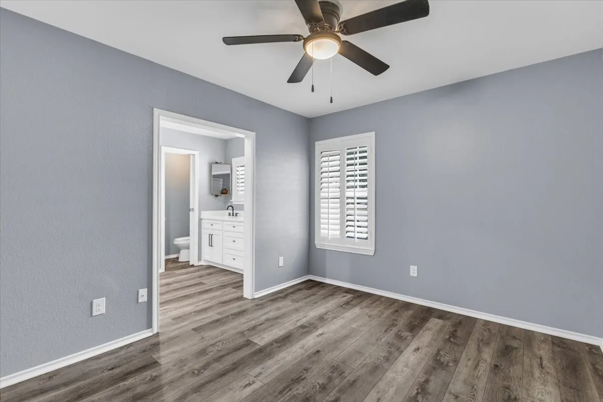This bright and spacious bedroom serves as an ideal first primary or mother-in-law suite, finished with neutral paint and continuous luxury vinyl plank flooring, and featuring a convenient attached ensuite bathroom, a modern lighted ceiling fan, and classic plantation shutters.