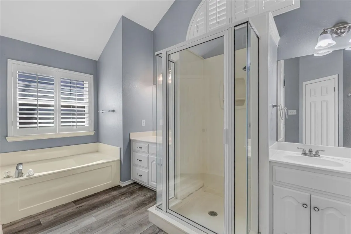 This bright, functional second primary bathroom features a dual-vanity layout with ample storage, a central walk-in shower, and a vaulted ceiling with an architectural arched window that maximizes natural light.