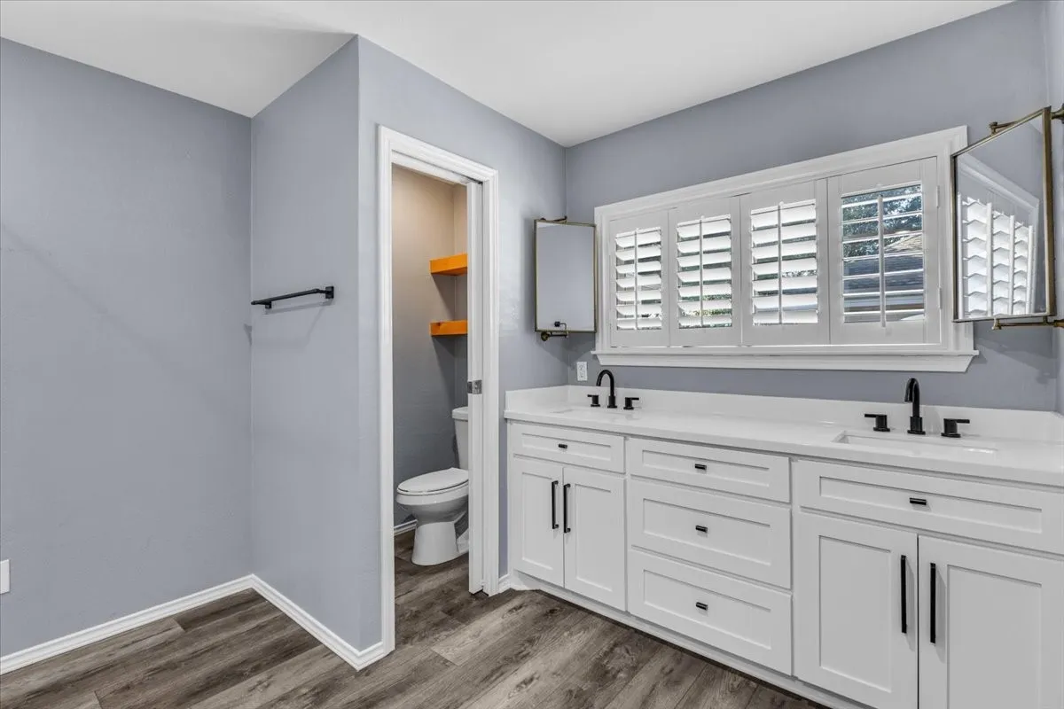 This alternate view of the primary bathroom highlights the expansive double vanity, offering abundant storage with crisp white cabinetry and striking matte black hardware. A large central window equipped with premium custom plantation shutters bathes the space in natural light, while dual pivot mirrors add a touch of elegance. The thoughtful layout is completed by a private water closet that features stylish built-in floating wood shelves.