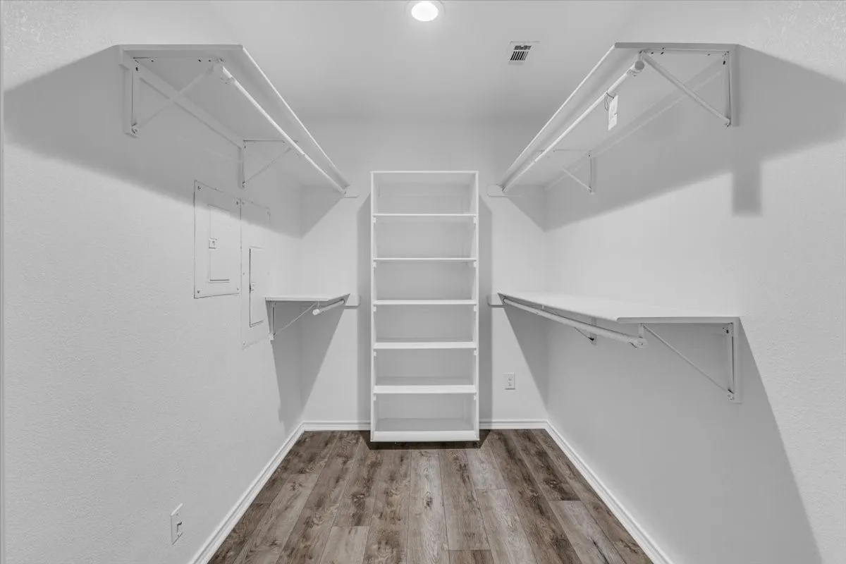 The first primary bedroom features a generously sized walk-in closet designed for optimal organization, complete with a built-in central shelving tower, ample hanging racks, bright overhead lighting, and beautiful continuous luxury vinyl flooring.