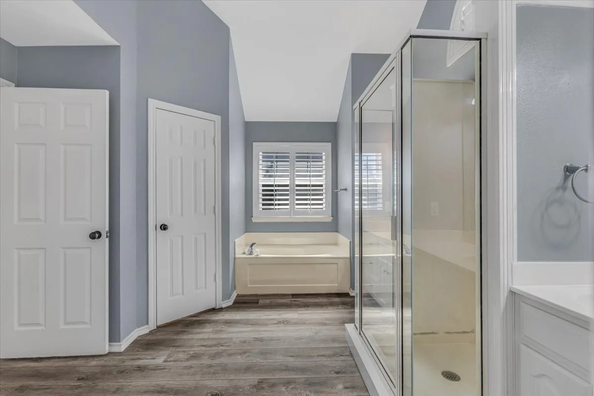 This expansive second primary bathroom offers a spa-like retreat, featuring a soothing soaking tub, a separate walk-in shower, dual vanities, and plantation shutters that perfectly balance privacy with natural light.