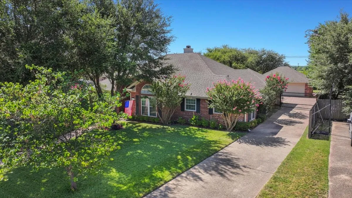 Showcasing exceptional curb appeal, this classic brick home is beautifully framed by mature shade trees, a well-manicured lawn, and vibrant blooming crepe myrtles. A long concrete driveway leads to a secure metal gate, offering both privacy and easy access to the detached garage in the rear. Elegant exterior details like a prominent arched front window and dark accent shutters complete this picturesque setting.